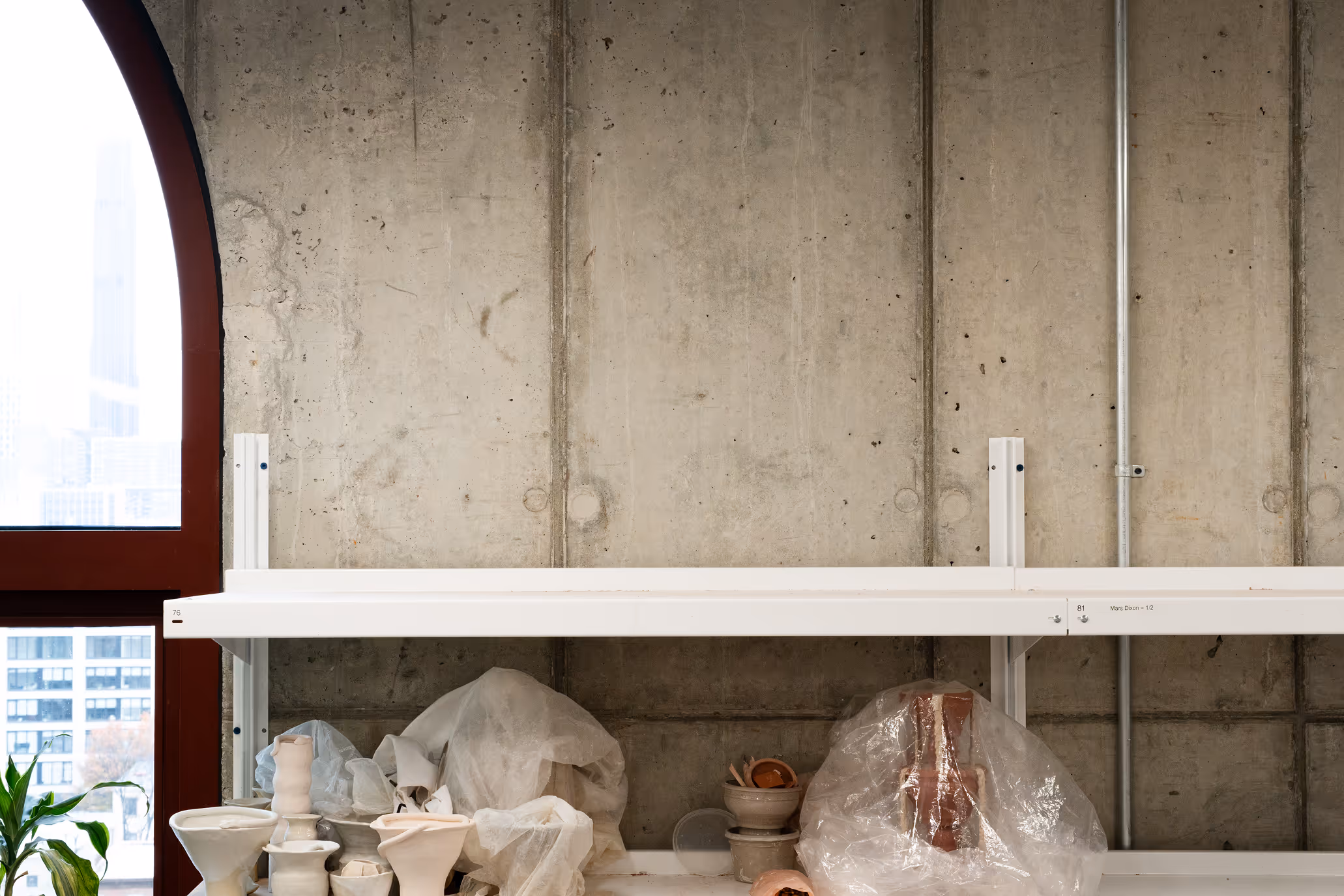 White shelf against a concrete wall holding various covered ceramic pottery pieces and a partially visible plant near a red window frame.