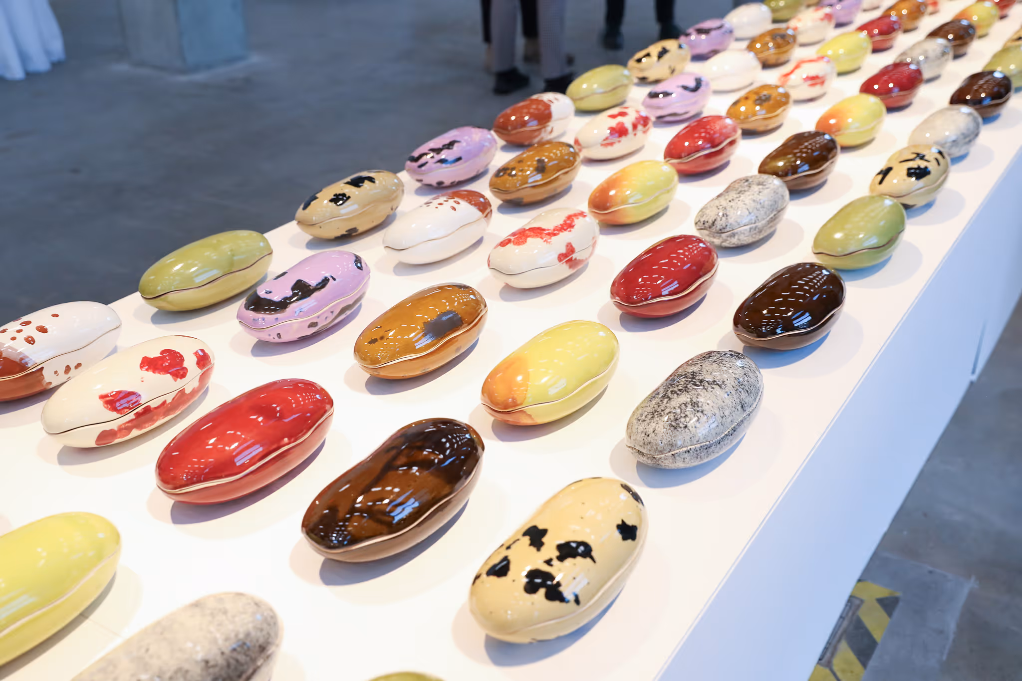 Rows of glossy, colorful ceramic beans with various patterns displayed on a white table in a gallery space.