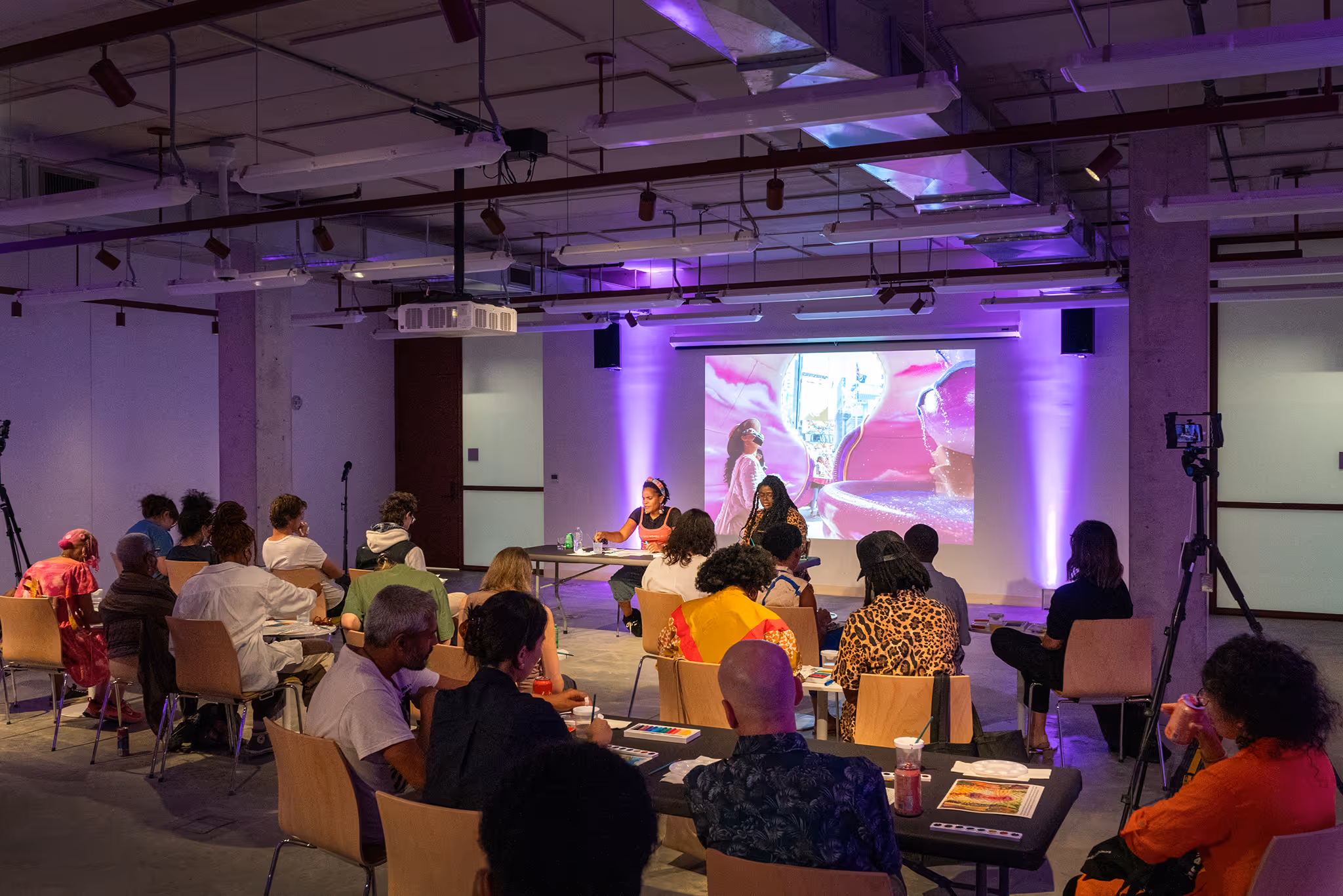 Audience seated in a dimly lit room watching a presentation with two speakers at a table and a colorful image projected on a screen.