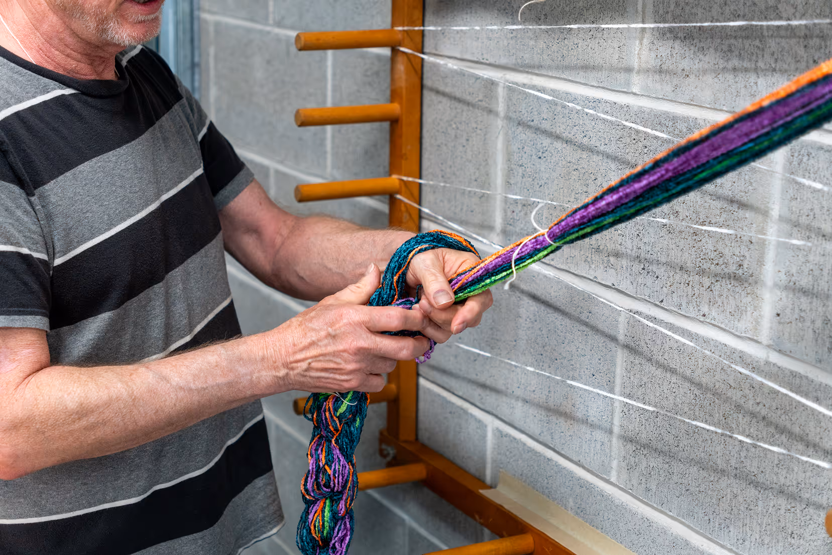 Man in striped shirt weaving colorful yarn on a wooden loom against a gray brick wall.