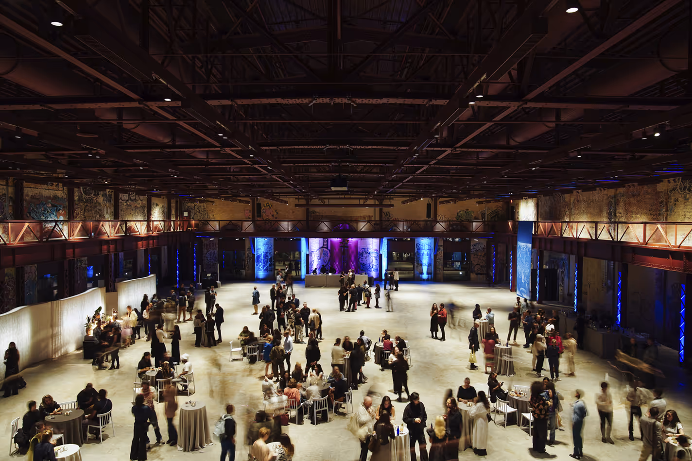 Large indoor event space with exposed ceiling beams, colorful uplighting, and groups of people mingling around tables.