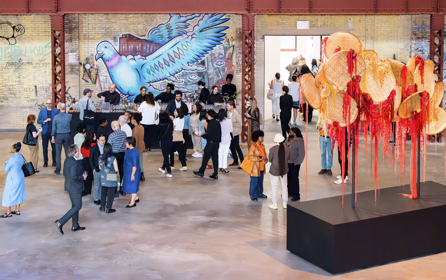 Art gallery event with diverse people mingling near a bar and a large hanging fiber art installation with red tassels.