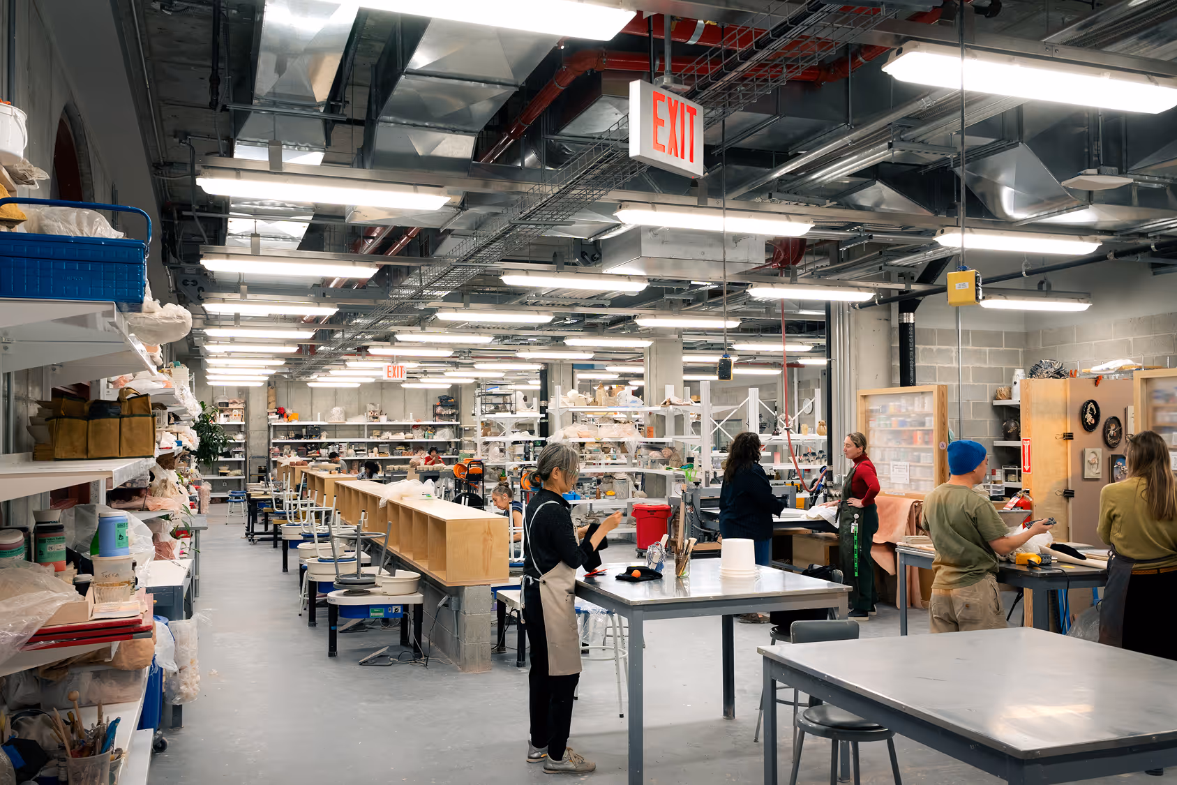 Busy ceramics studio with people working at tables surrounded by shelves filled with pottery supplies and finished pieces.