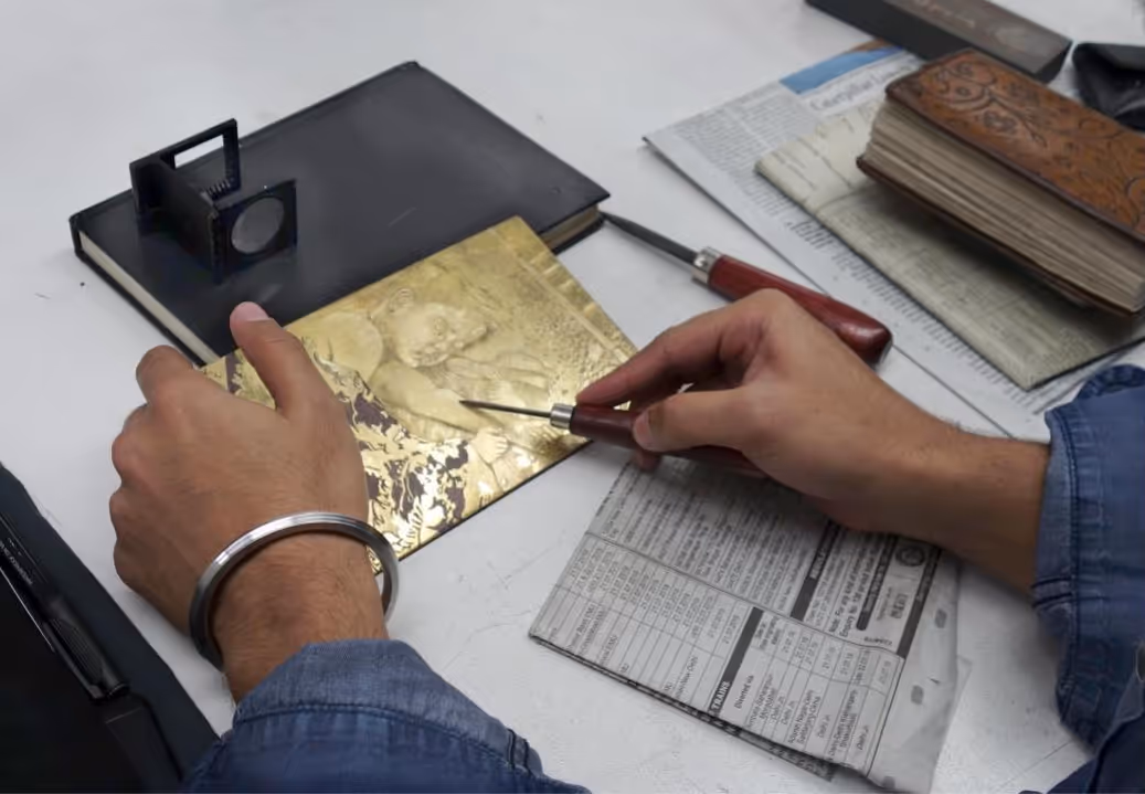 Person engraving a detailed figure on a gold metal plate using a precision tool on a cluttered work desk.