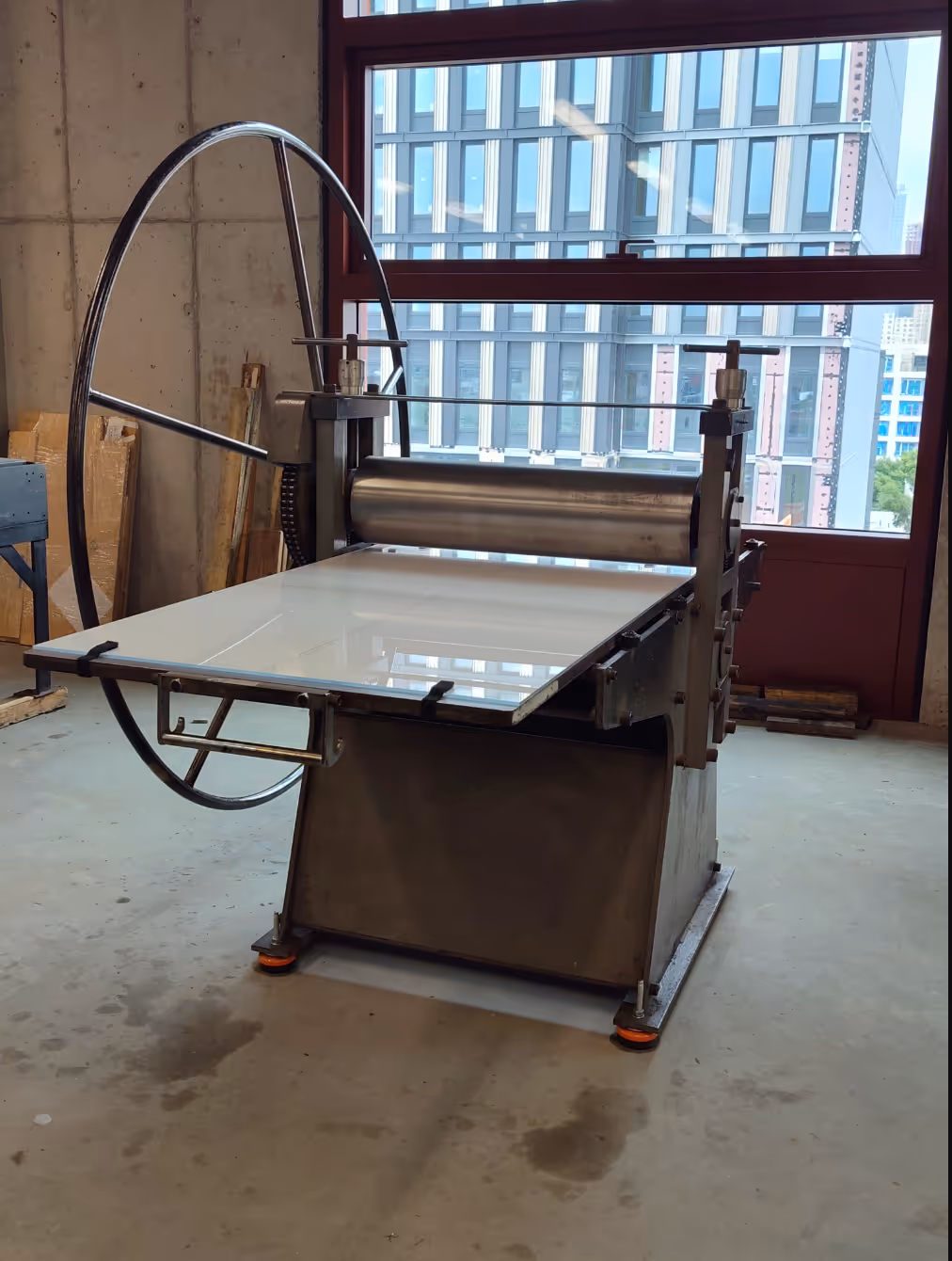 Large industrial printing press with a metal roller and a hand wheel in a workshop with concrete floor and windows showing a modern building outside.