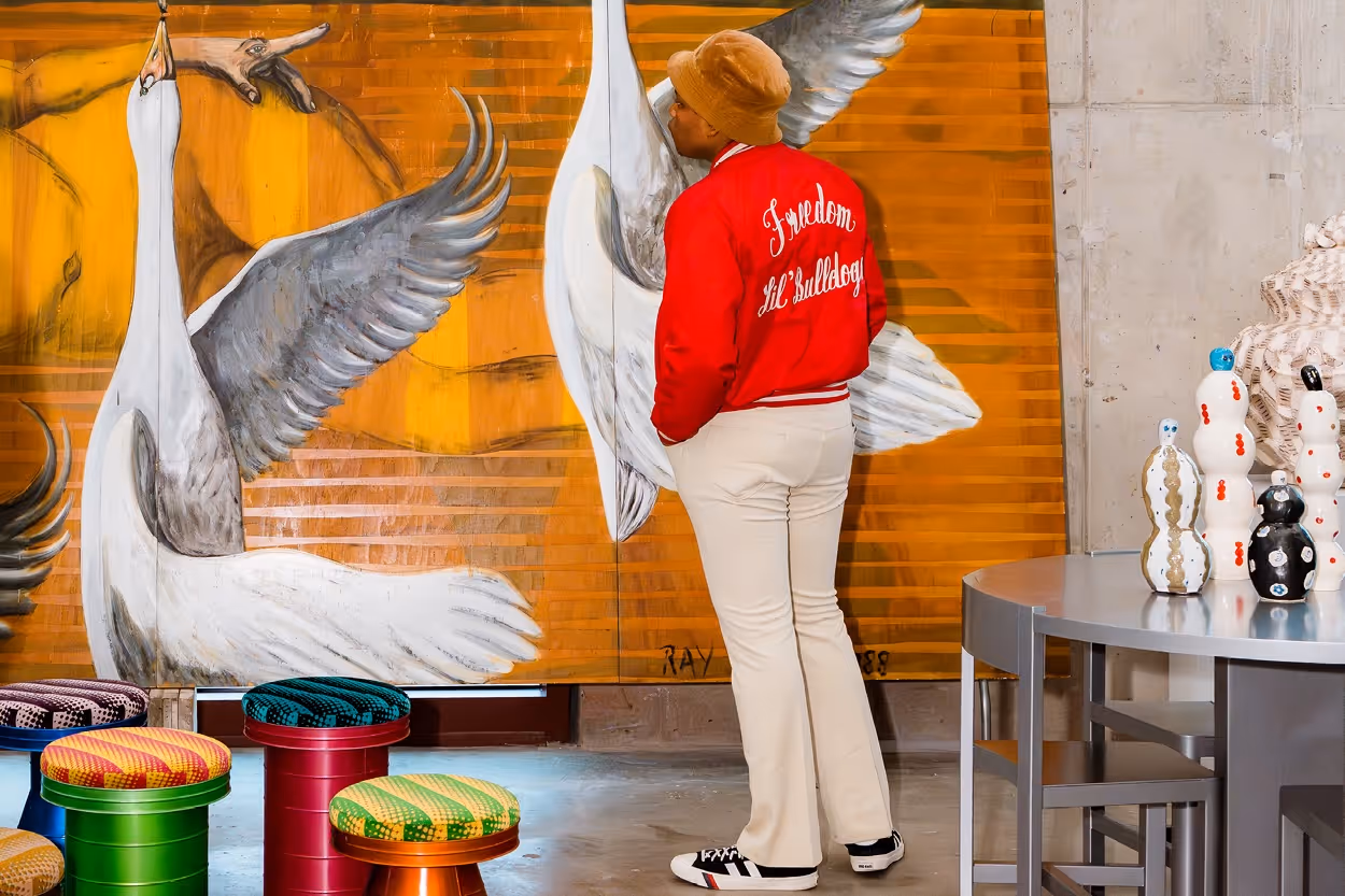 Person in a red jacket and beige pants leaning forward to look closely at large bird paintings on an orange wall in a modern art space.