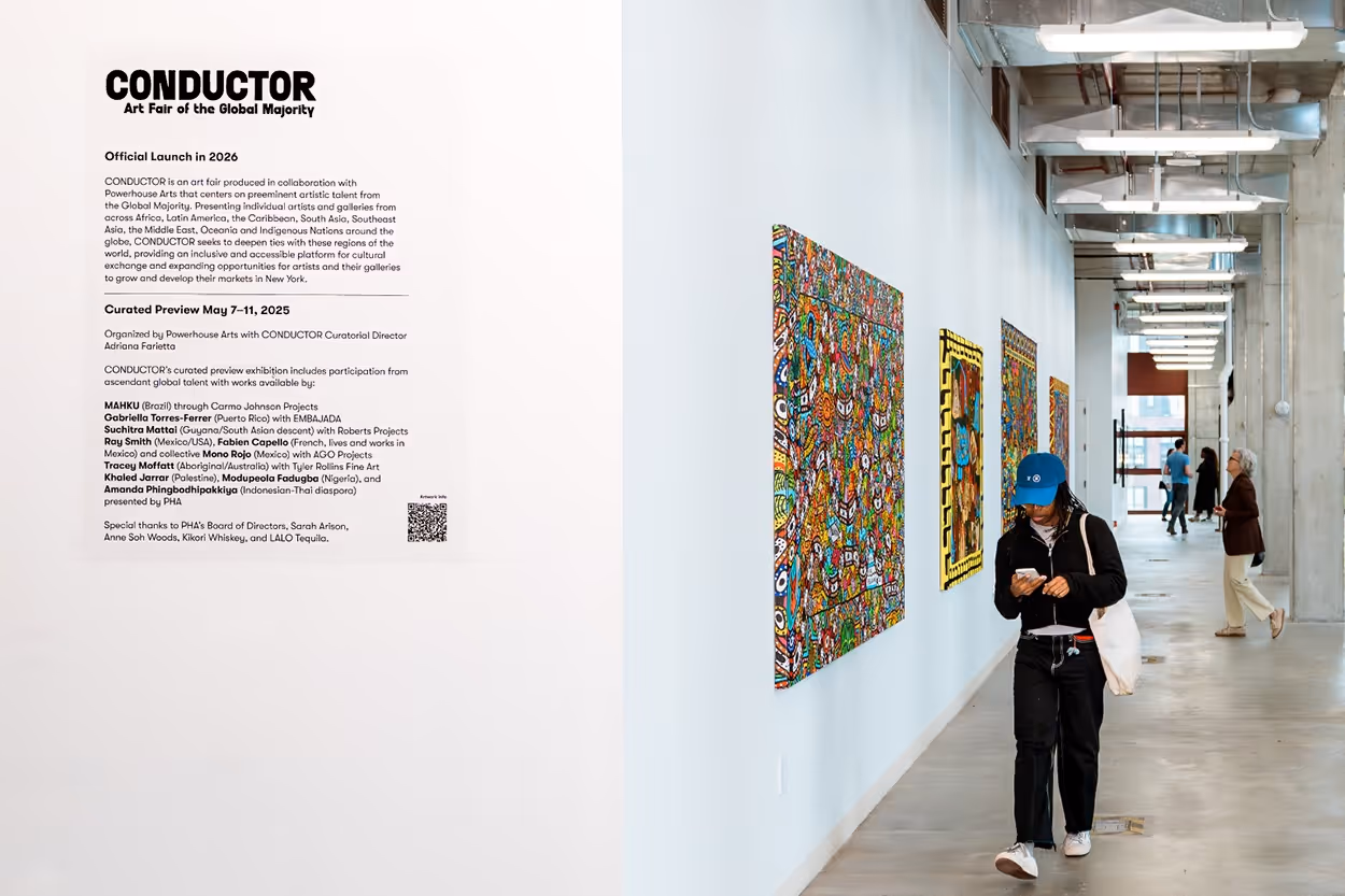 Art gallery hallway with colorful paintings on the wall and people walking, with a large white sign detailing the Conductor Art Fair launch and curated preview.