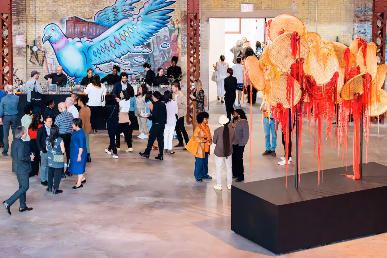 People mingling at an art gallery with a colorful mural of a large blue bird on the wall and a woven art installation with red hanging threads on a black pedestal.