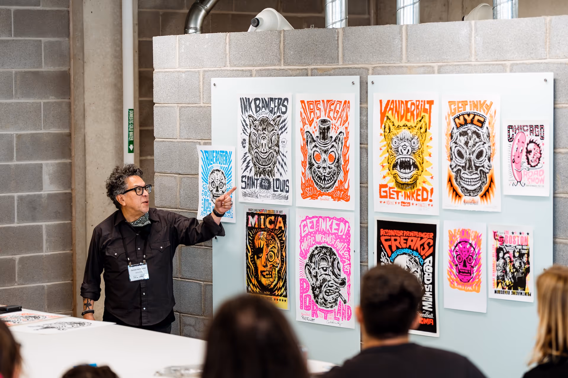 Man with glasses pointing at colorful graphic posters on a wall during a presentation with audience in foreground.