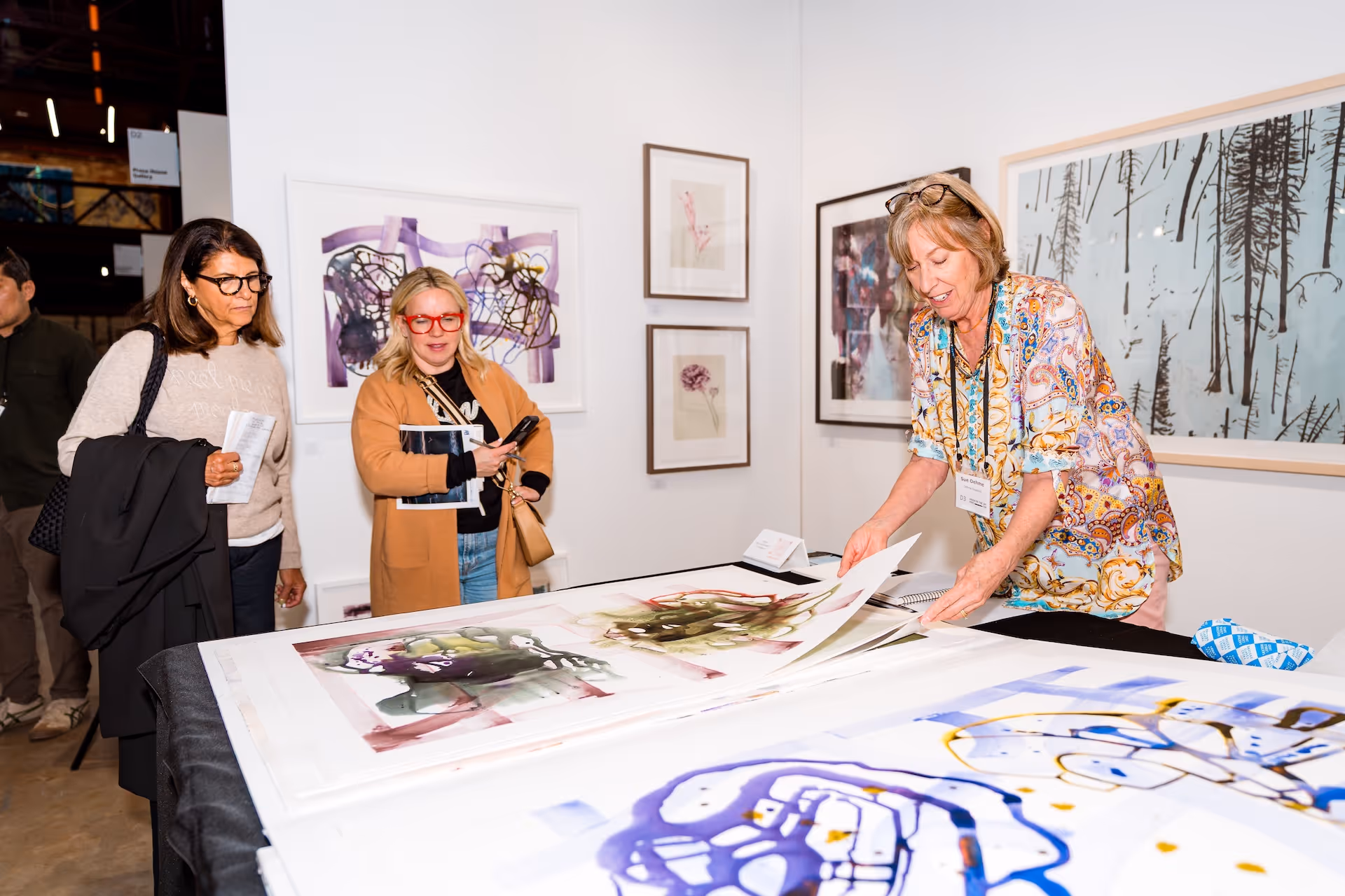 Woman showing and flipping through abstract art prints on a table to two women at an art gallery or fair.