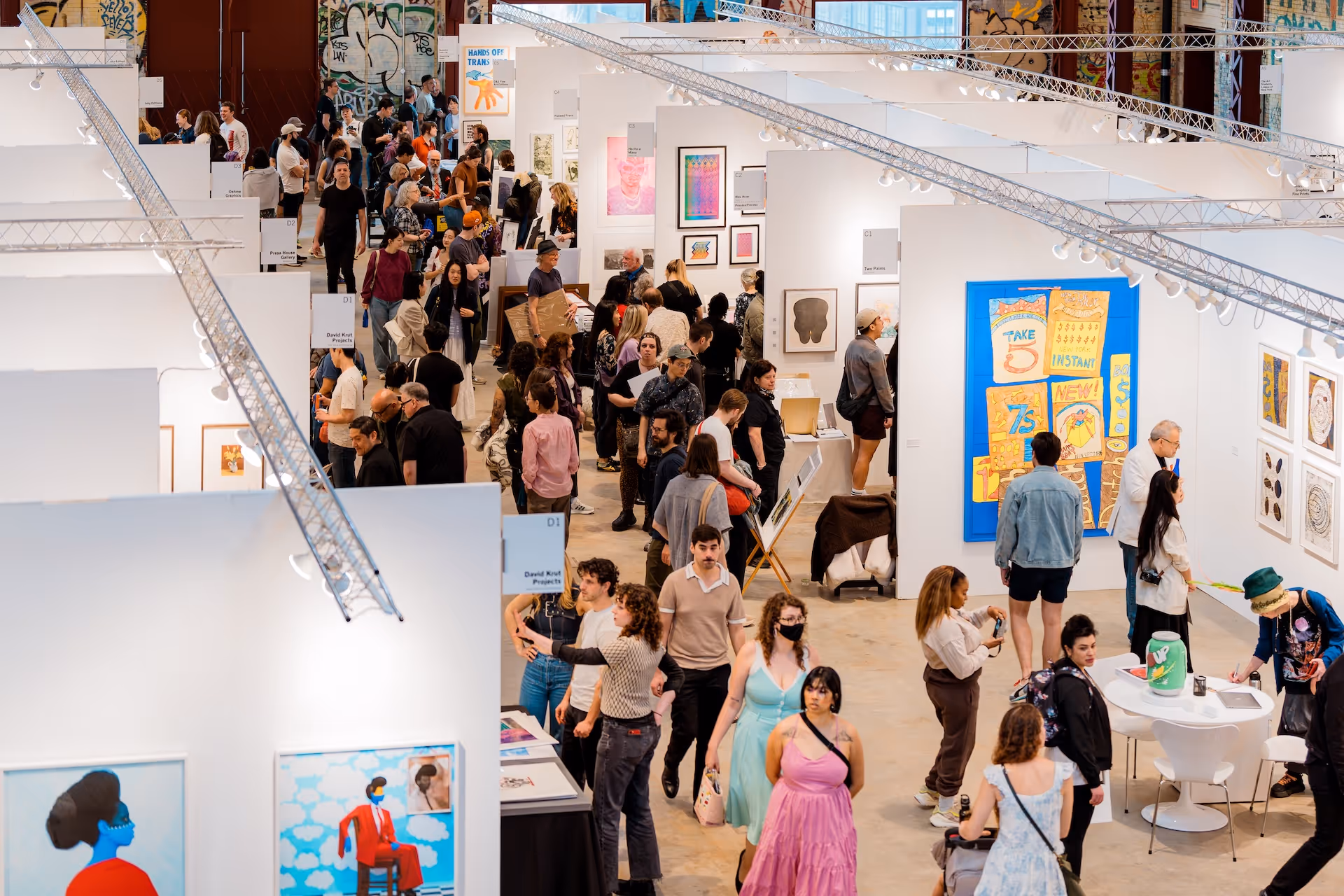 Crowded indoor art fair with many people viewing colorful artworks displayed on white walls.