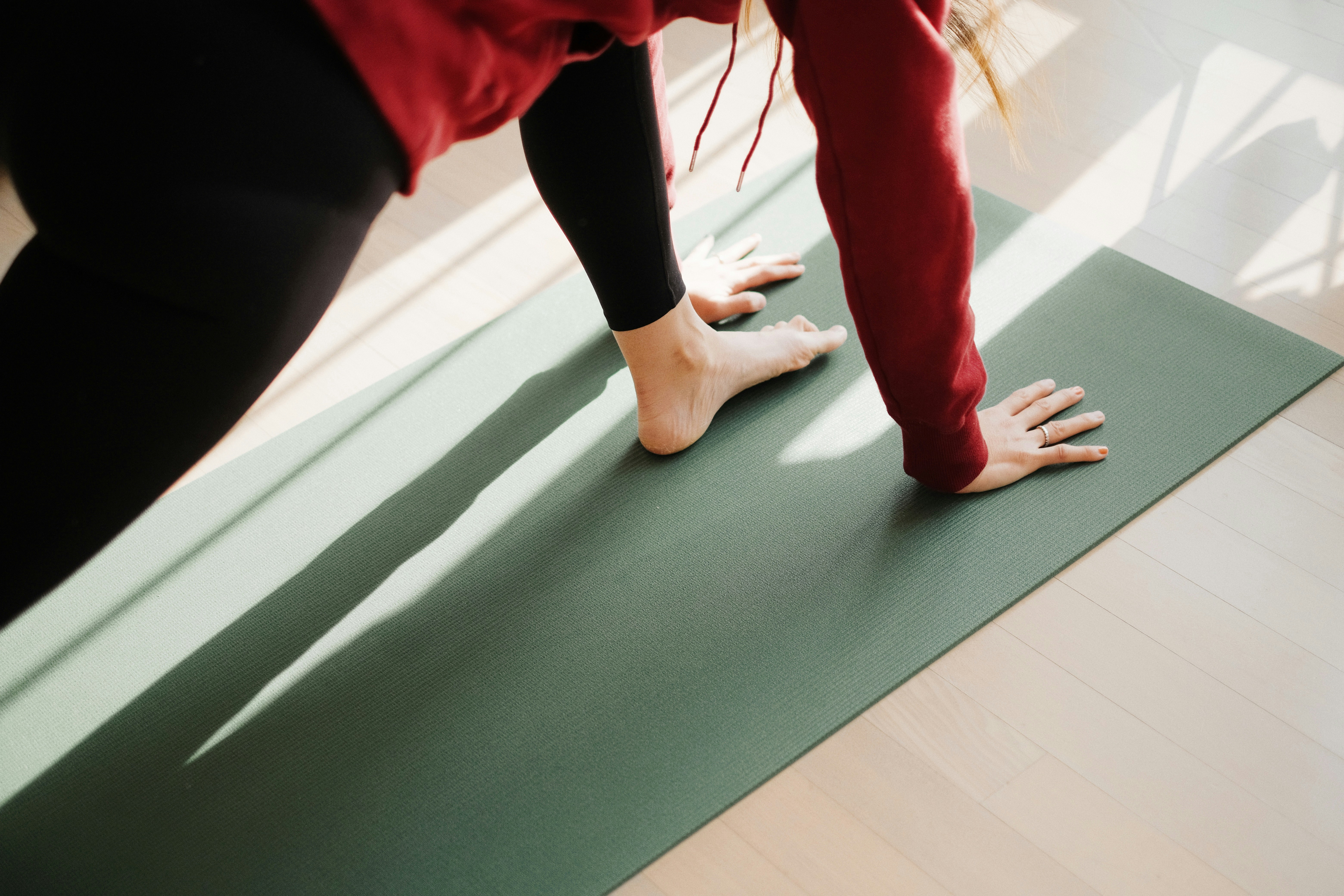Une femme est allongée sur le dos sur un tapis de yoga en jute tressé, dans la pose de Savasana. Ses bras sont étendus le long de son corps, paumes vers le haut, et ses yeux sont fermés. Elle porte un débardeur et un legging gris clair. En arrière-plan, il y a une fenêtre avec des rideaux blancs transparents et une plante verte en pot.