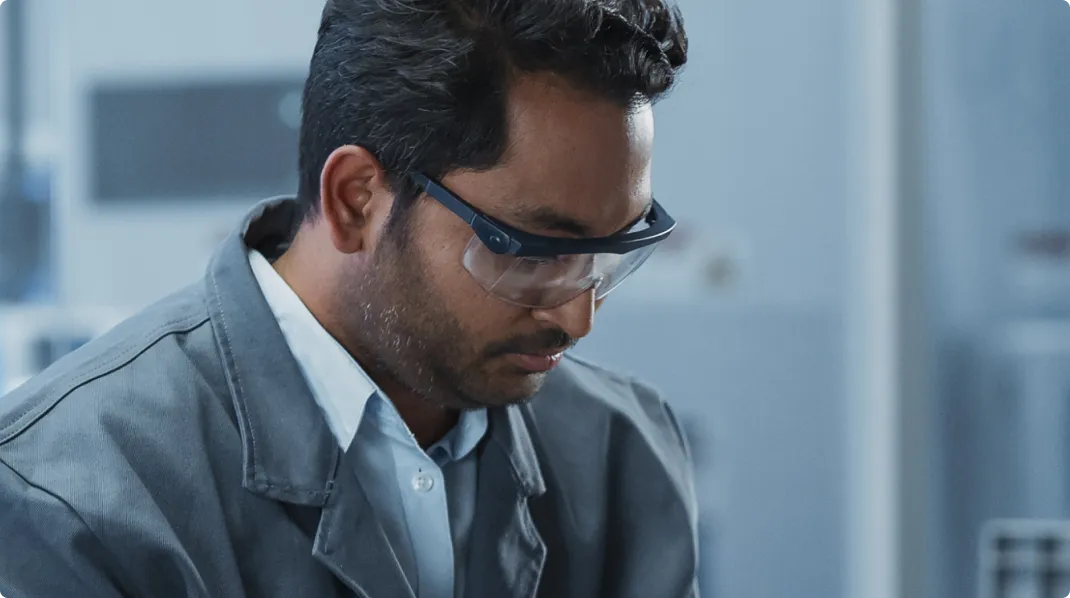 Man wearing safety glasses and a gray jacket, focused on his work in a lab or industrial setting.
