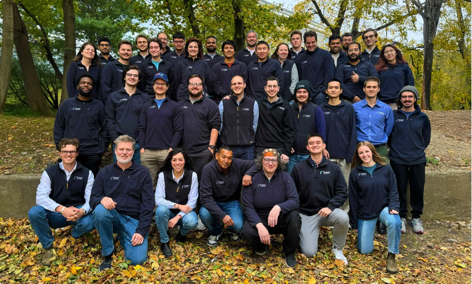 Large group of 34 people posing outside in a park with autumn leaves on the ground, many wearing dark jackets with Tutor logos.