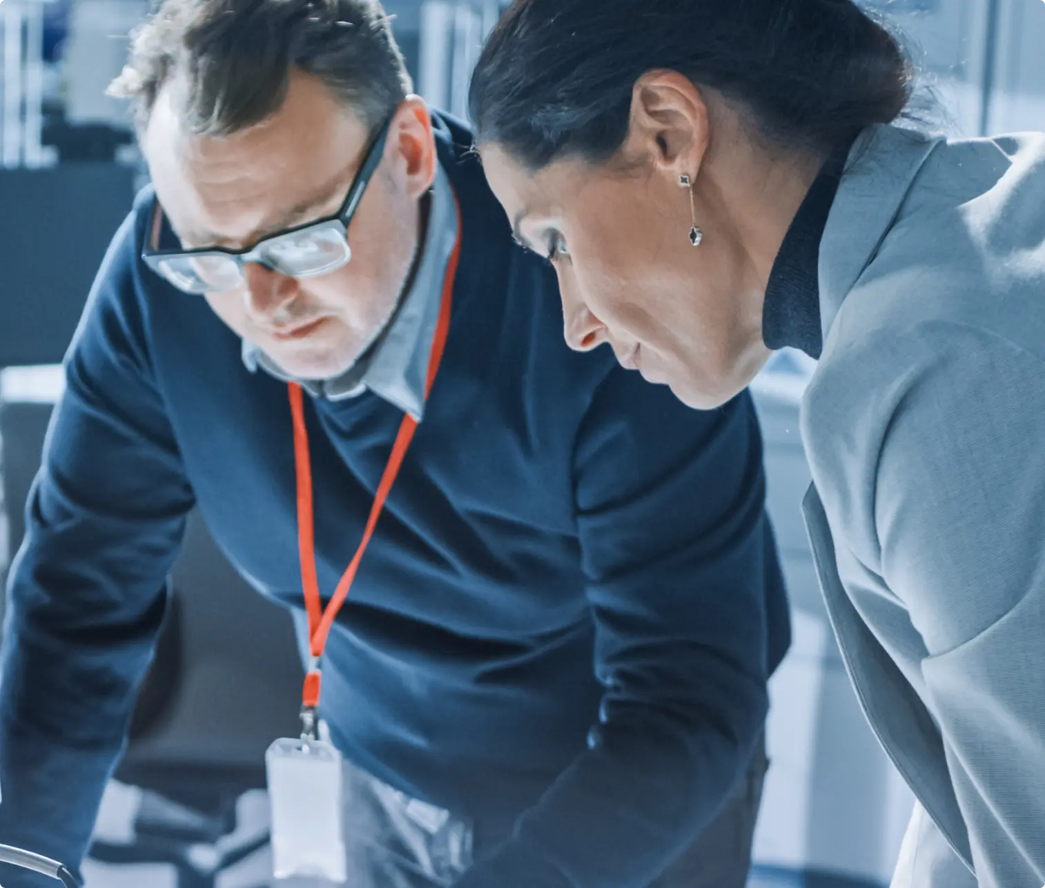 Man in glasses and navy sweater with red lanyard and woman in gray blazer closely reviewing documents together.