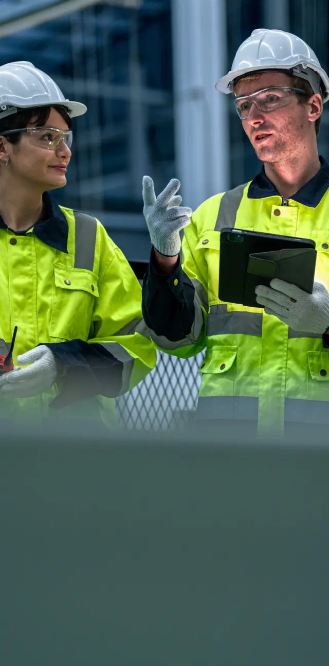 Two construction workers in high-visibility jackets, hard hats, gloves, and safety glasses discussing work, one holding a tablet.