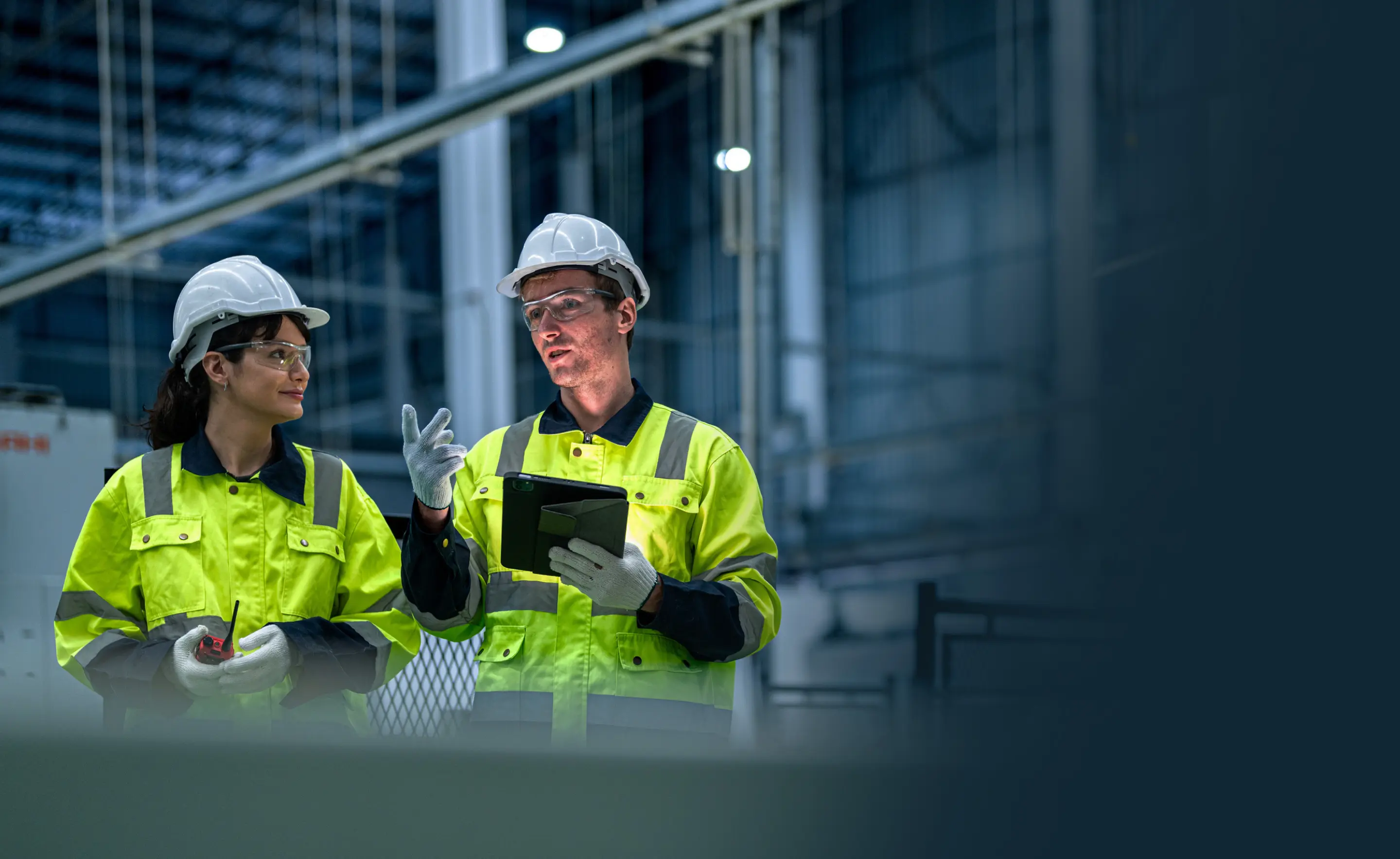Two construction workers in high-visibility jackets, hard hats, gloves, and safety glasses discussing work, one holding a tablet.