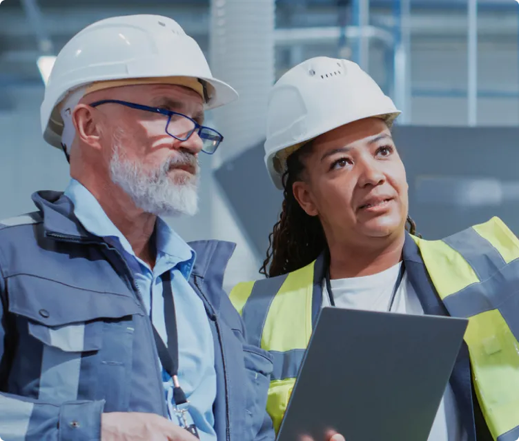 Two construction workers wearing white hard hats and safety vests, one holding a tablet, engaged in discussion inside an industrial building.