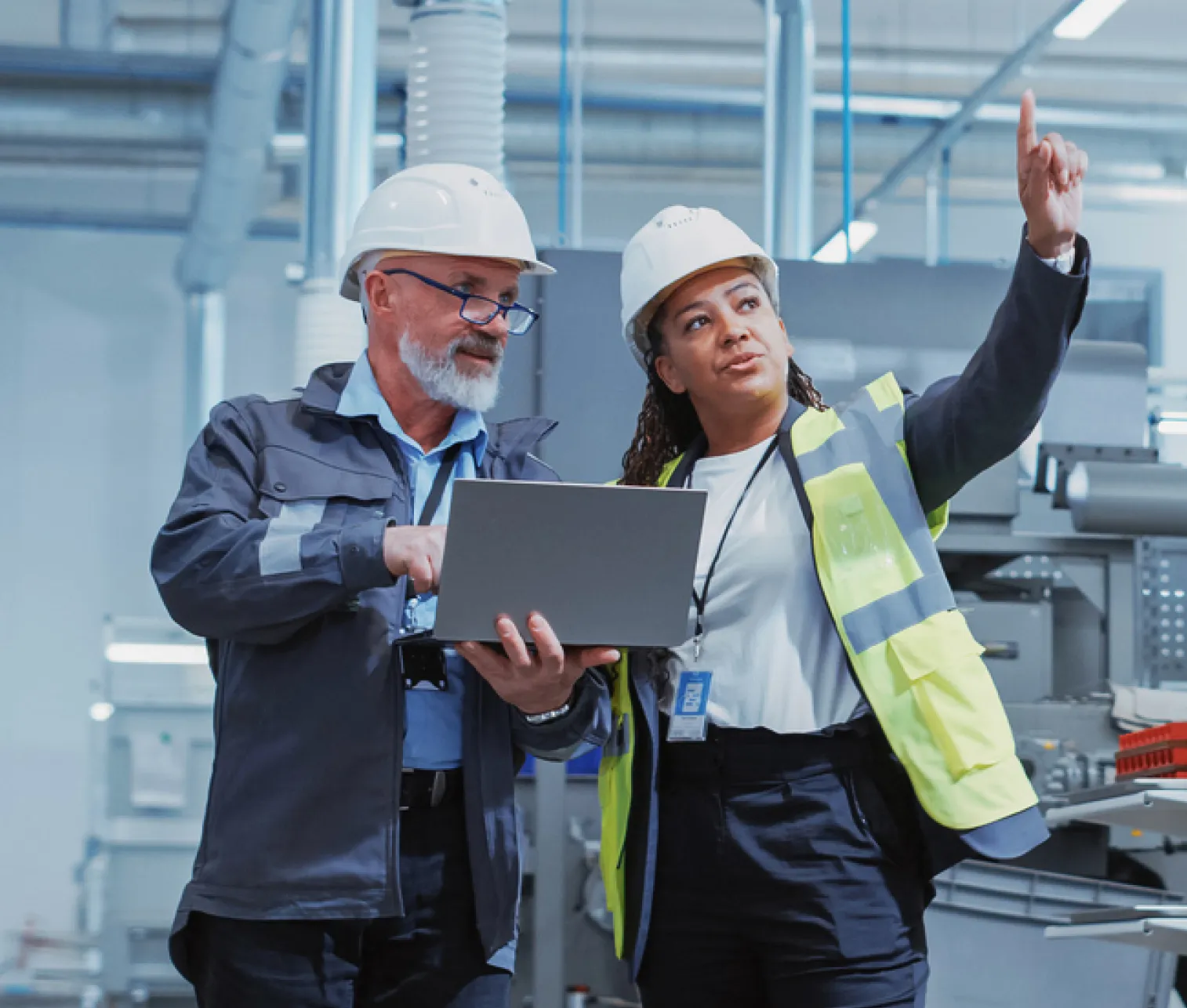 Two engineers in a factory wearing hard hats, one holding a laptop while the other points towards something.
