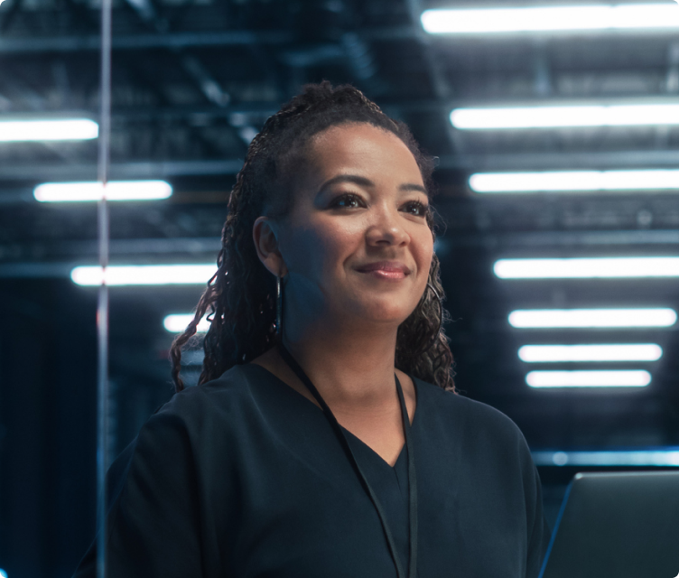 Smiling woman with curly hair wearing a dark top and lanyard in a modern office with bright overhead lights.