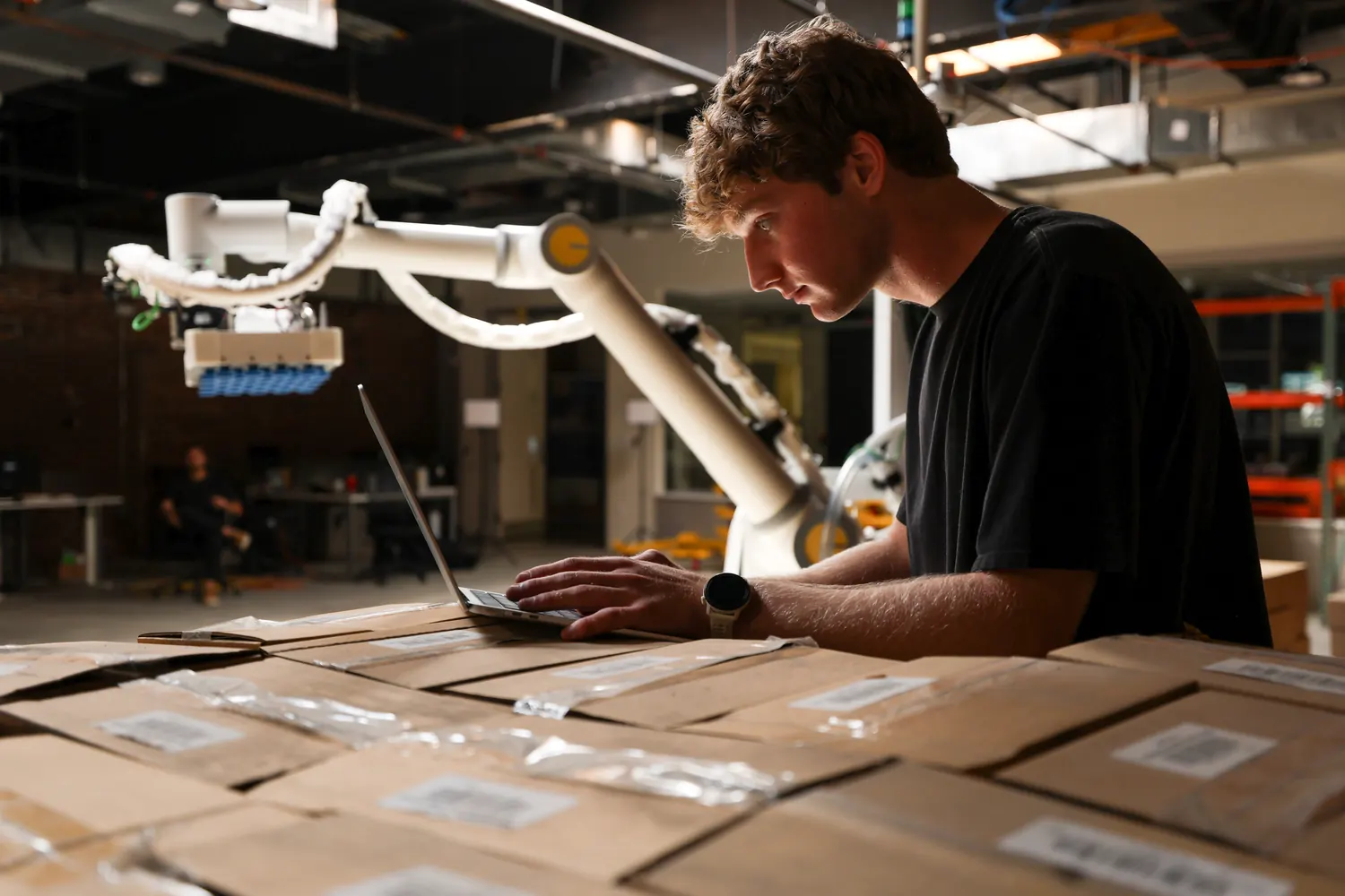 An engineer works on his laptop, which sits on top of a pallet of cases, with a Tutor Cassie in the background.