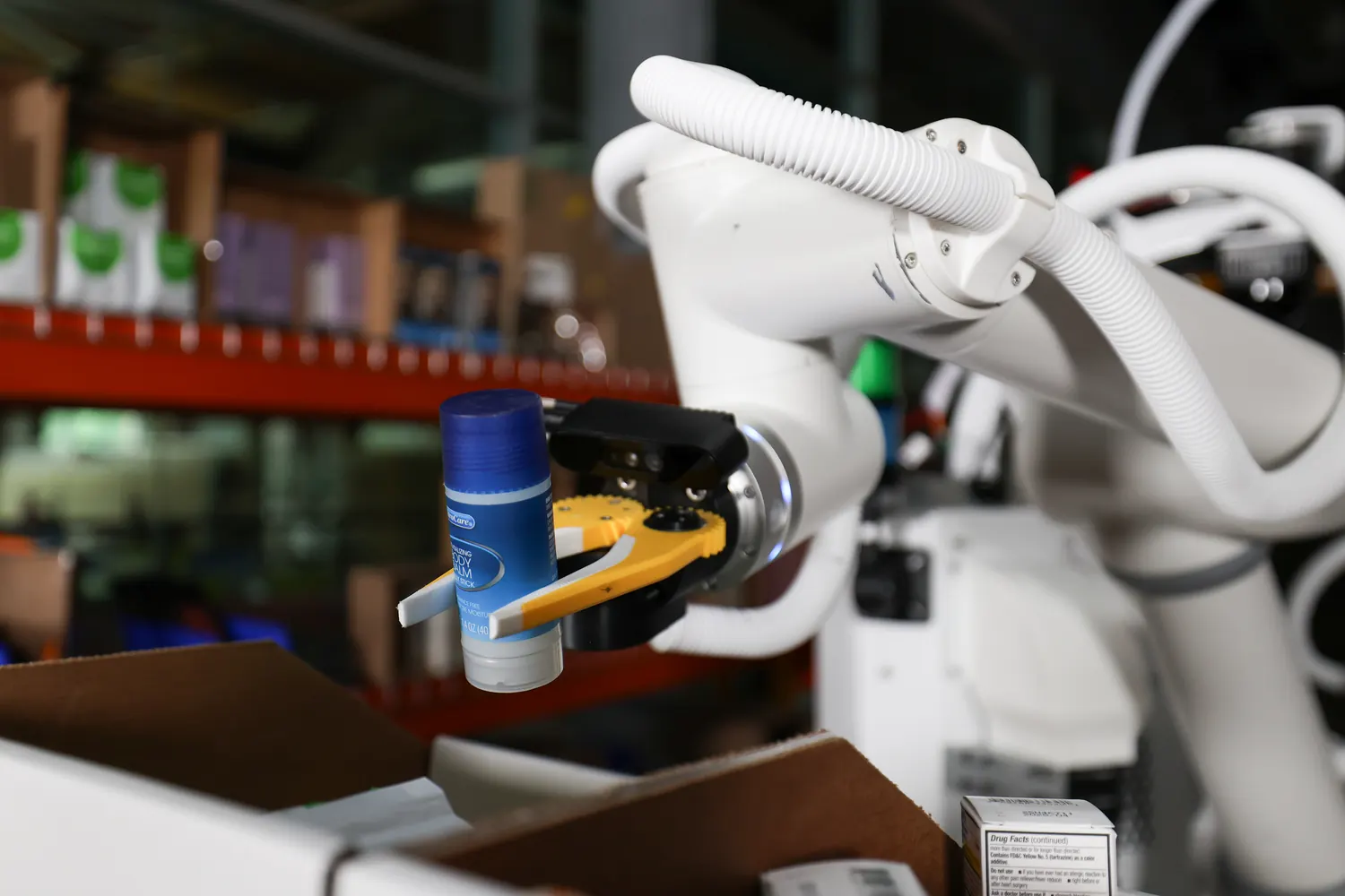Tutor robot Sonny's gripper holding a body balm stick above an open carton of retail inventory.
