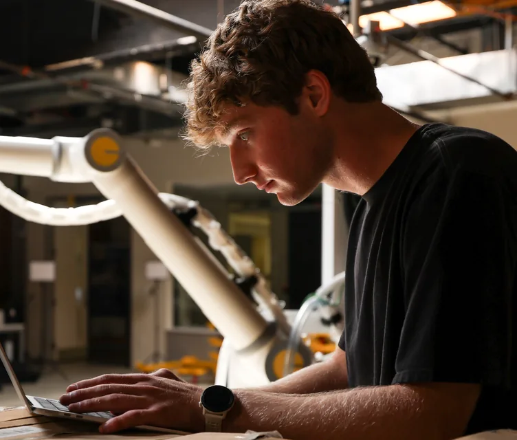 A Tutor engineer working on a laptop beside a robot arm on the factory floor.
