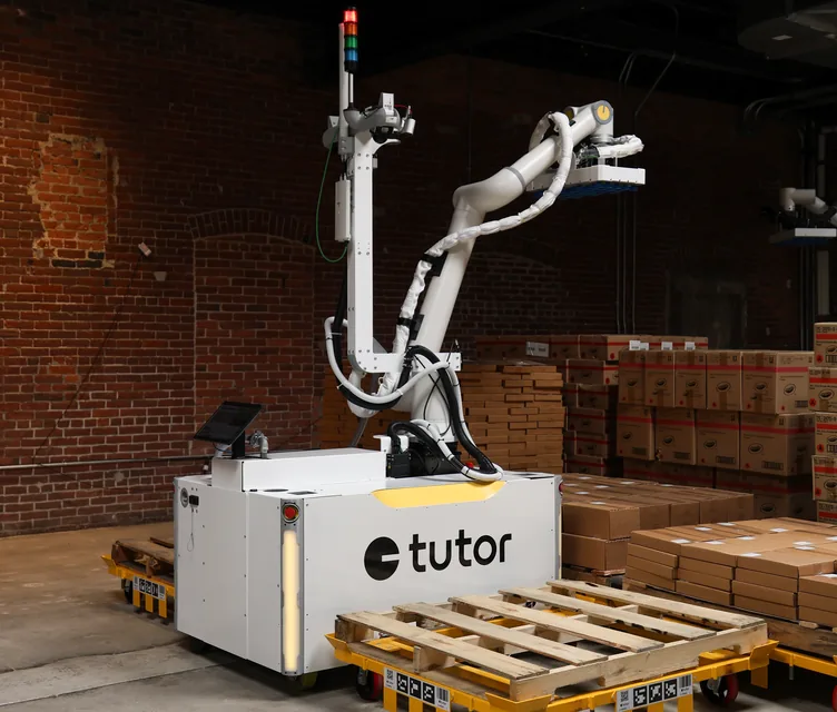 A Tutor Cassie robot alongside a pallet dolly and stacked cases in a warehouse.