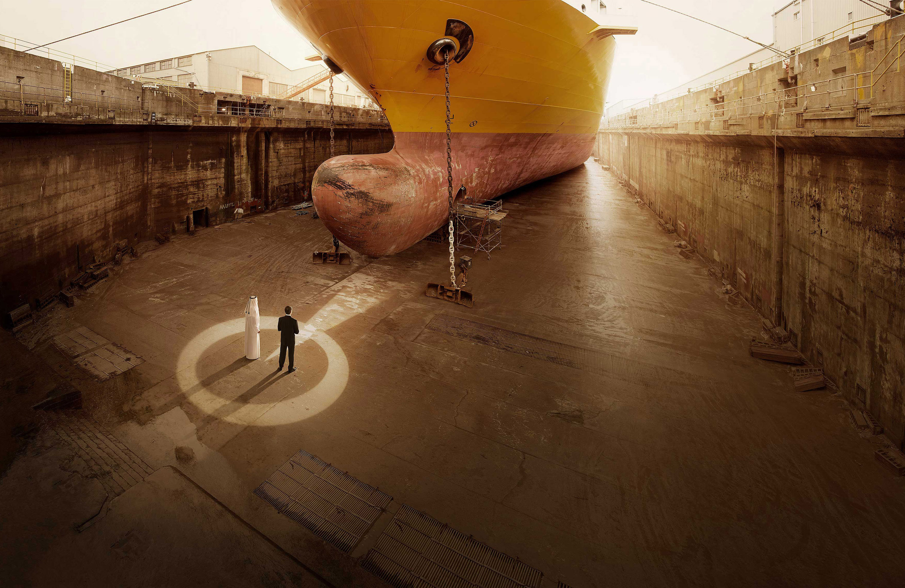Two men, one in a white robe and the other in a dark suit, standing in a dry dock next to the bow of a large ship.