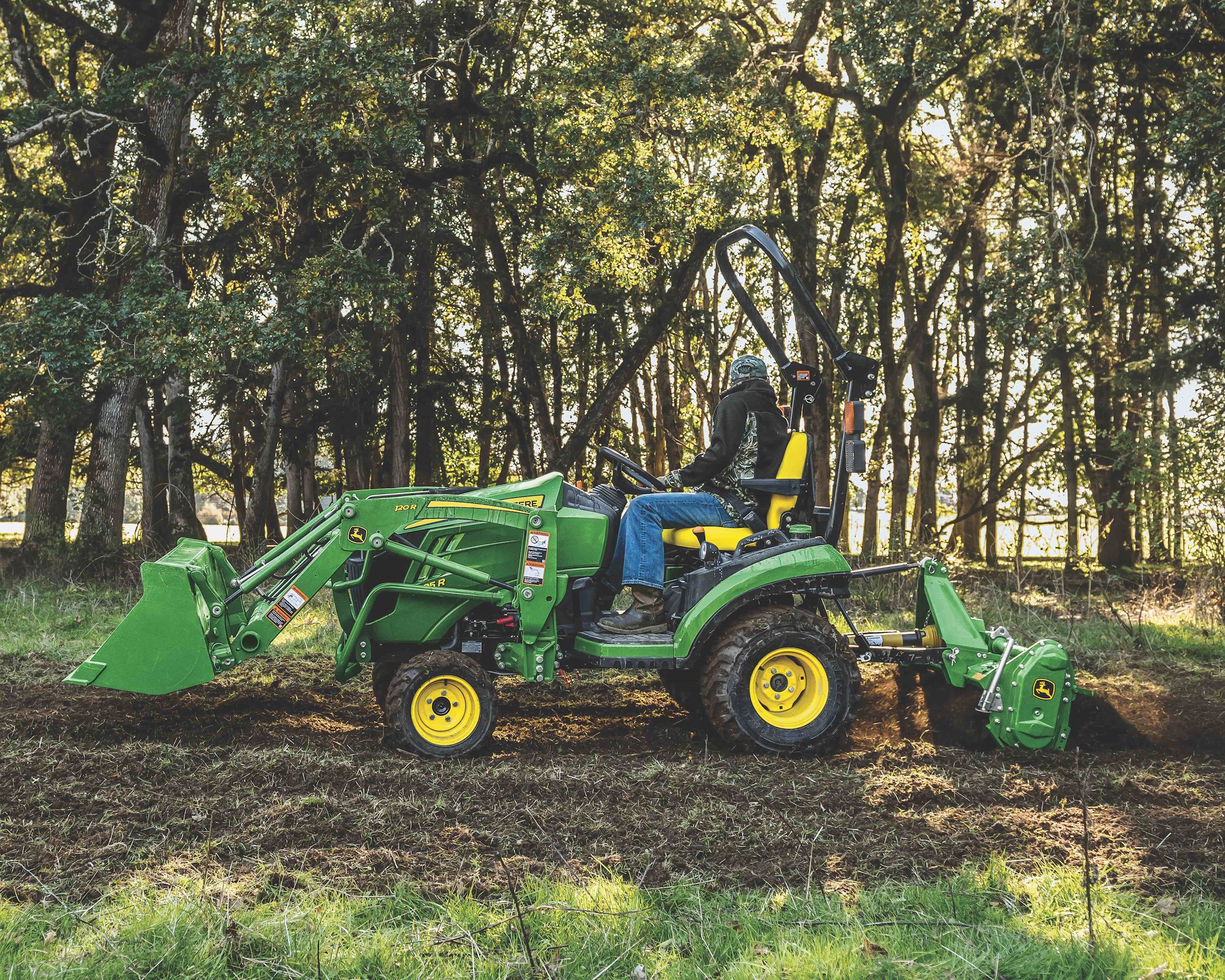 A John Deere 1025R tractor does work surrounded by trees in the woods.