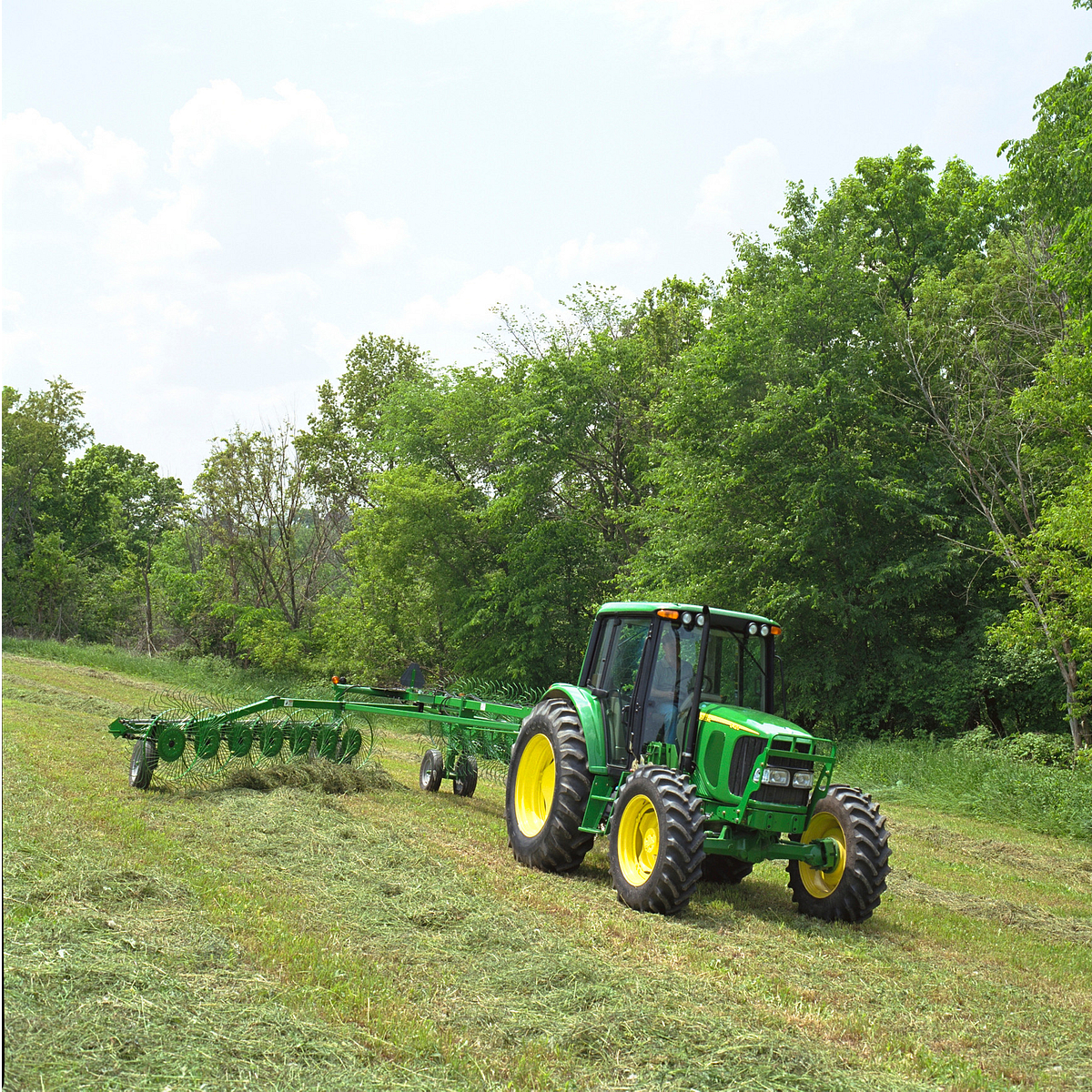 Front right, angled view of a tractor pulling a WR High-Capacity Wheel Rake across a hay field bordered by trees
