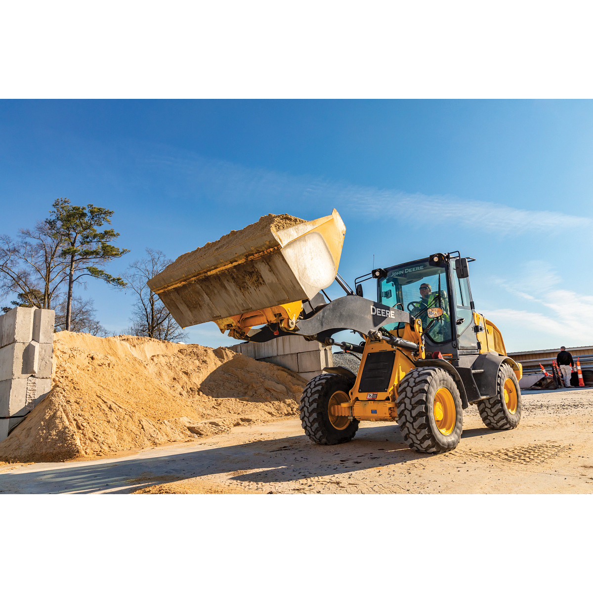 A wheel loader lifting a full bucket of sand at a construction site, with piled material, concrete blocks, trees, and a clear blue sky in the background.