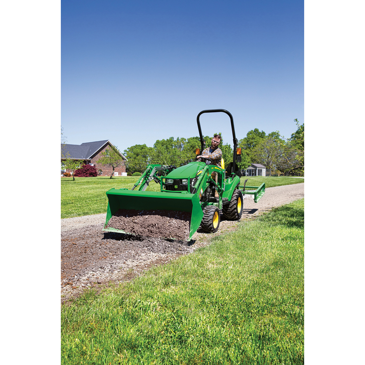 A person operates a small green tractor with a front loader attachment, leveling and spreading gravel along a driveway beside a grassy lawn, with houses and trees visible under a clear blue sky