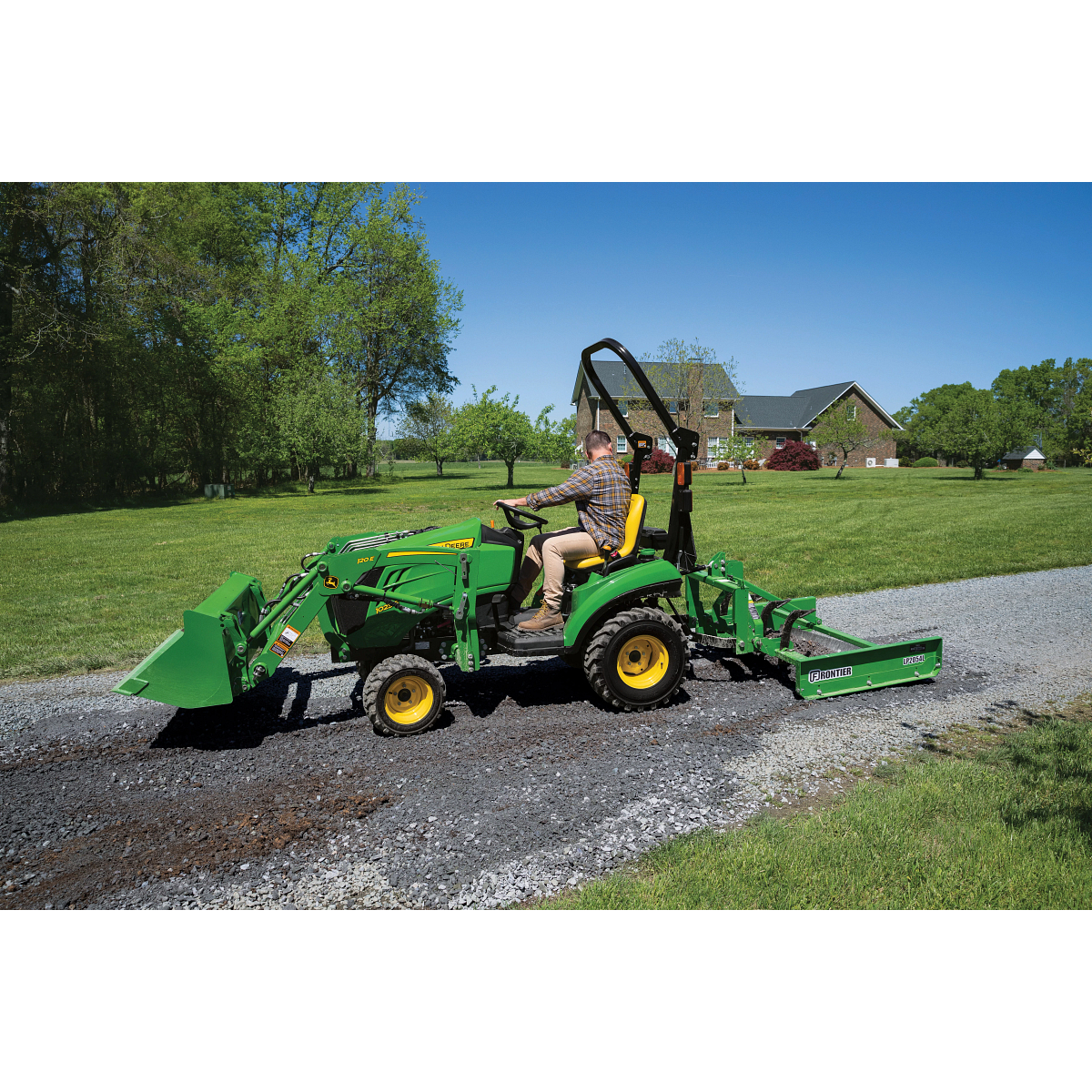 A person drives a small green tractor with a front loader and rear land plane implement along a gravel driveway on a sunny day, with grassy lawn and a brick house in the background