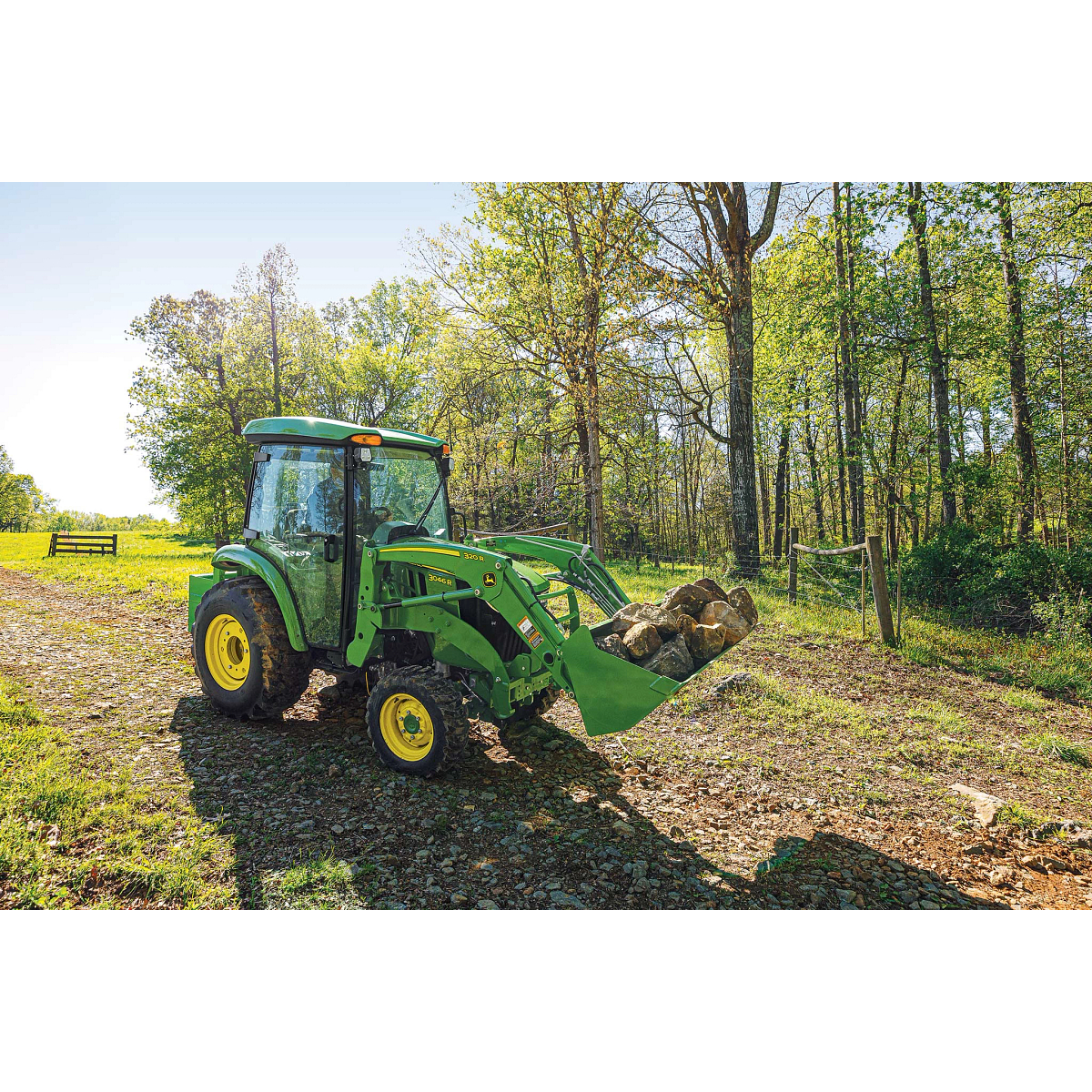 Front right view of a 3046R compact utility tractor with front-attached 320R loader hauling boulders across a dirt path near the woods