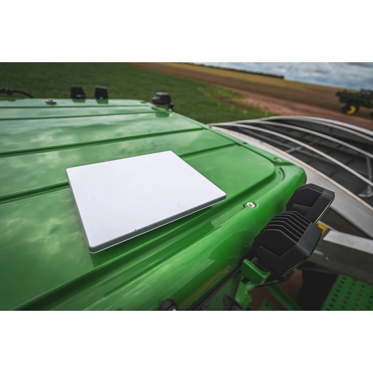 JDLink Boost Mini Terminal mounted on the roof of a green John Deere machine with attachments and field visible in the background.