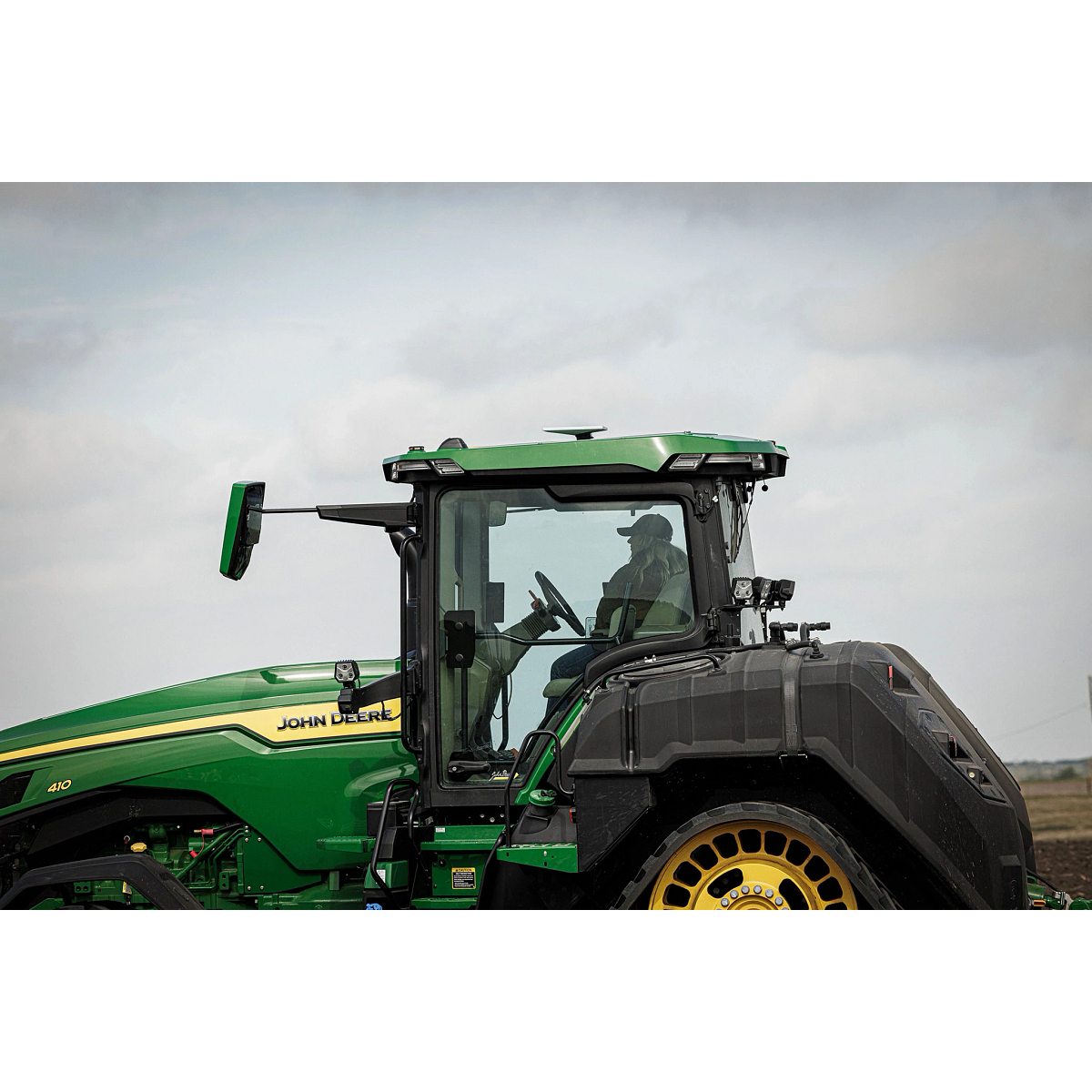 Side view of a John Deere 8R 410 tractor in a field, showing a roof-mounted JDLink Boost Mini Terminal above the cab with an operator seated inside.