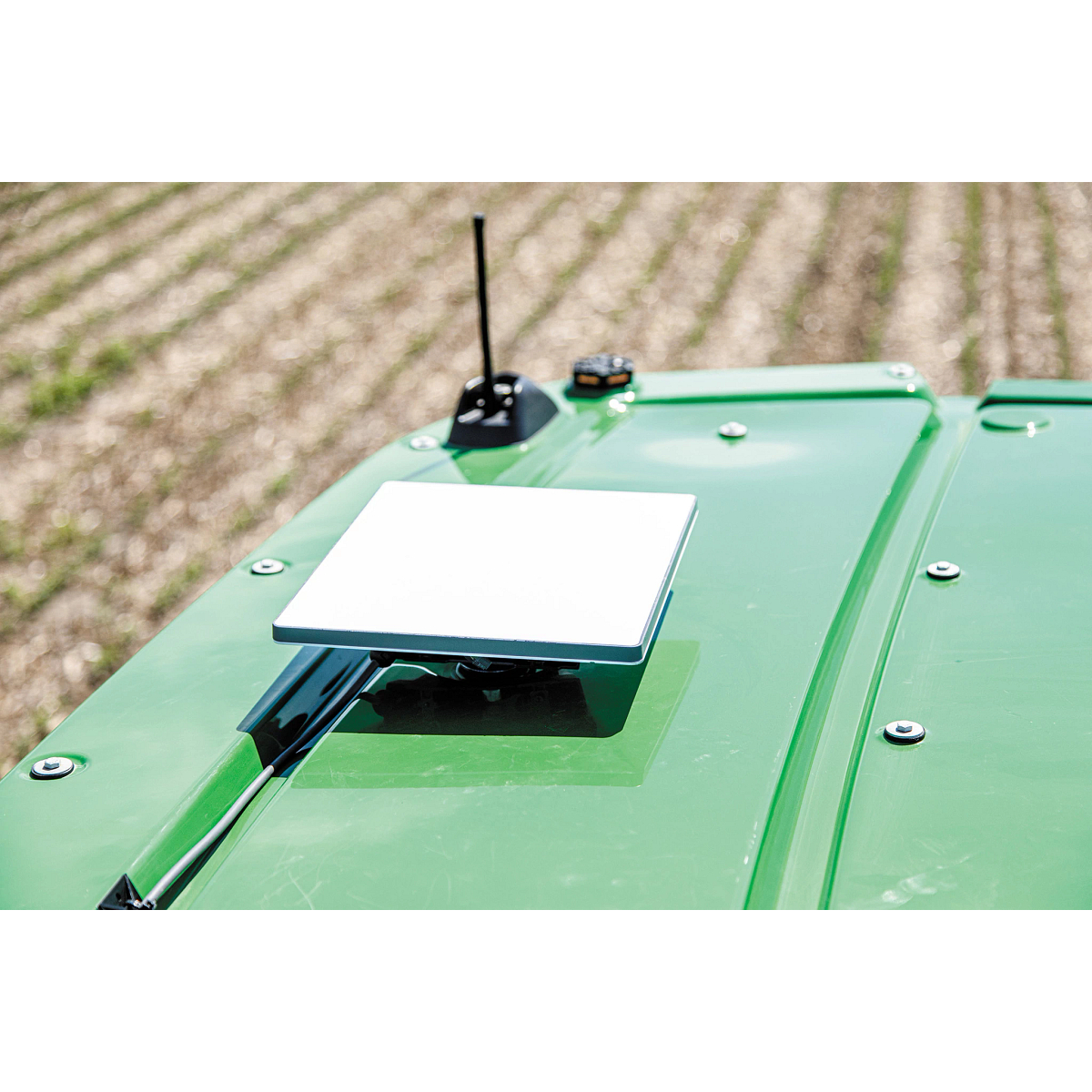 Close-up of a JDLink Boost Mini Terminal mounted on the roof of a John Deere machine near an antenna, with crop rows in the background.
