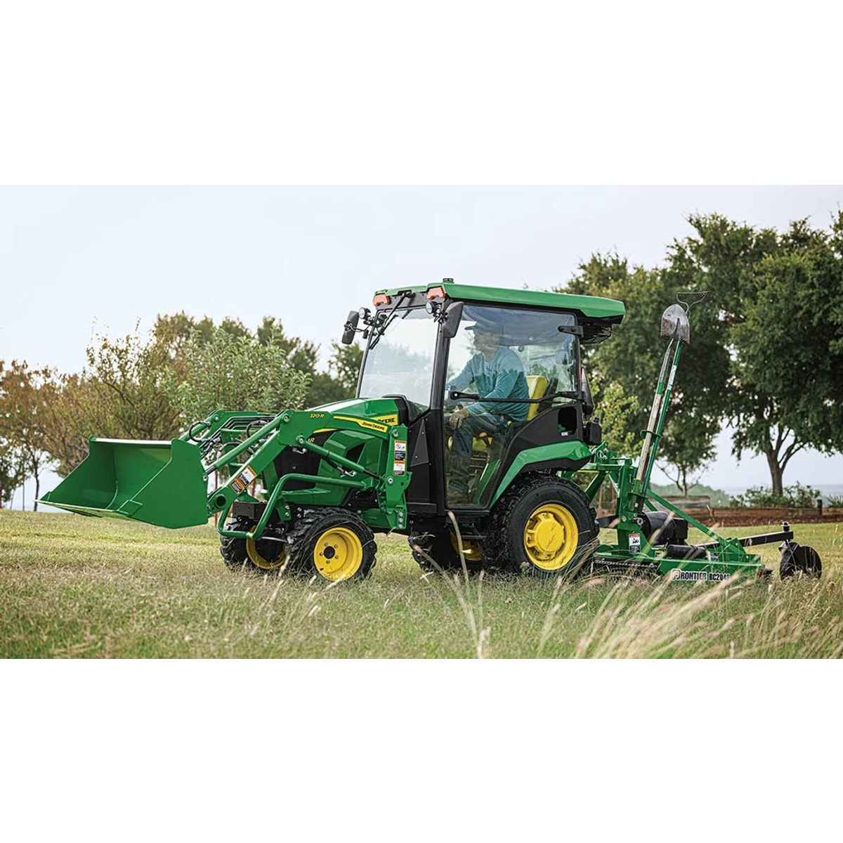 A cabbed green compact tractor with a loader pulls a rotary cutter over some grass.