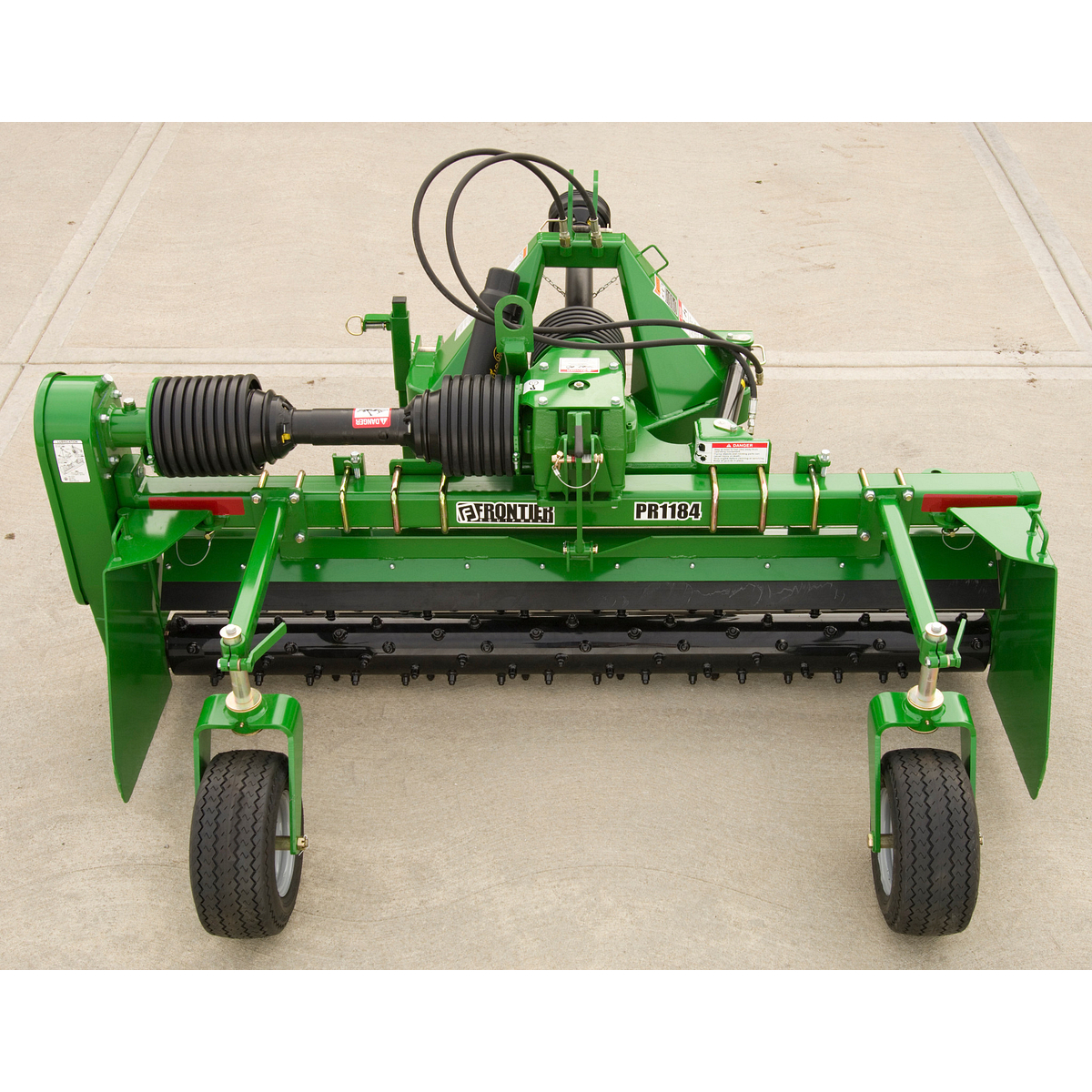 Frontier PR1184 rotary tiller attachment on concrete, featuring a wide cutting deck, central gearbox with PTO shaft, rows of blades underneath, and two support wheels.