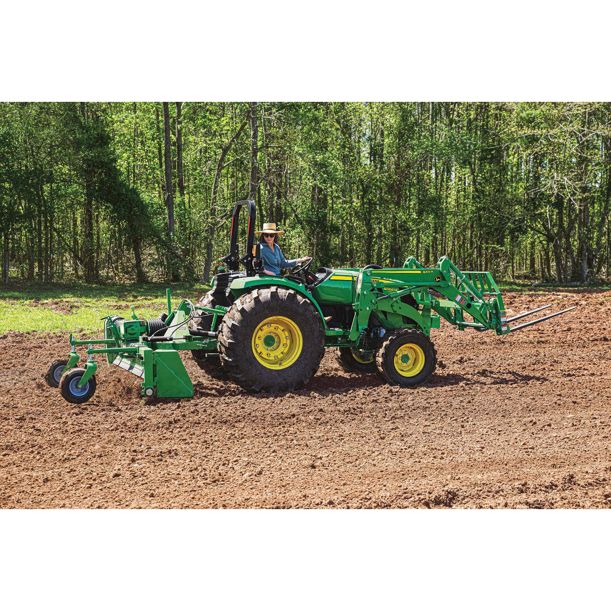Tractor with a front loader and tilling attachment drives across a brown, freshly tilled field. A person in a hat operates the tractor, with a line of leafy trees in the background under bright daylight.