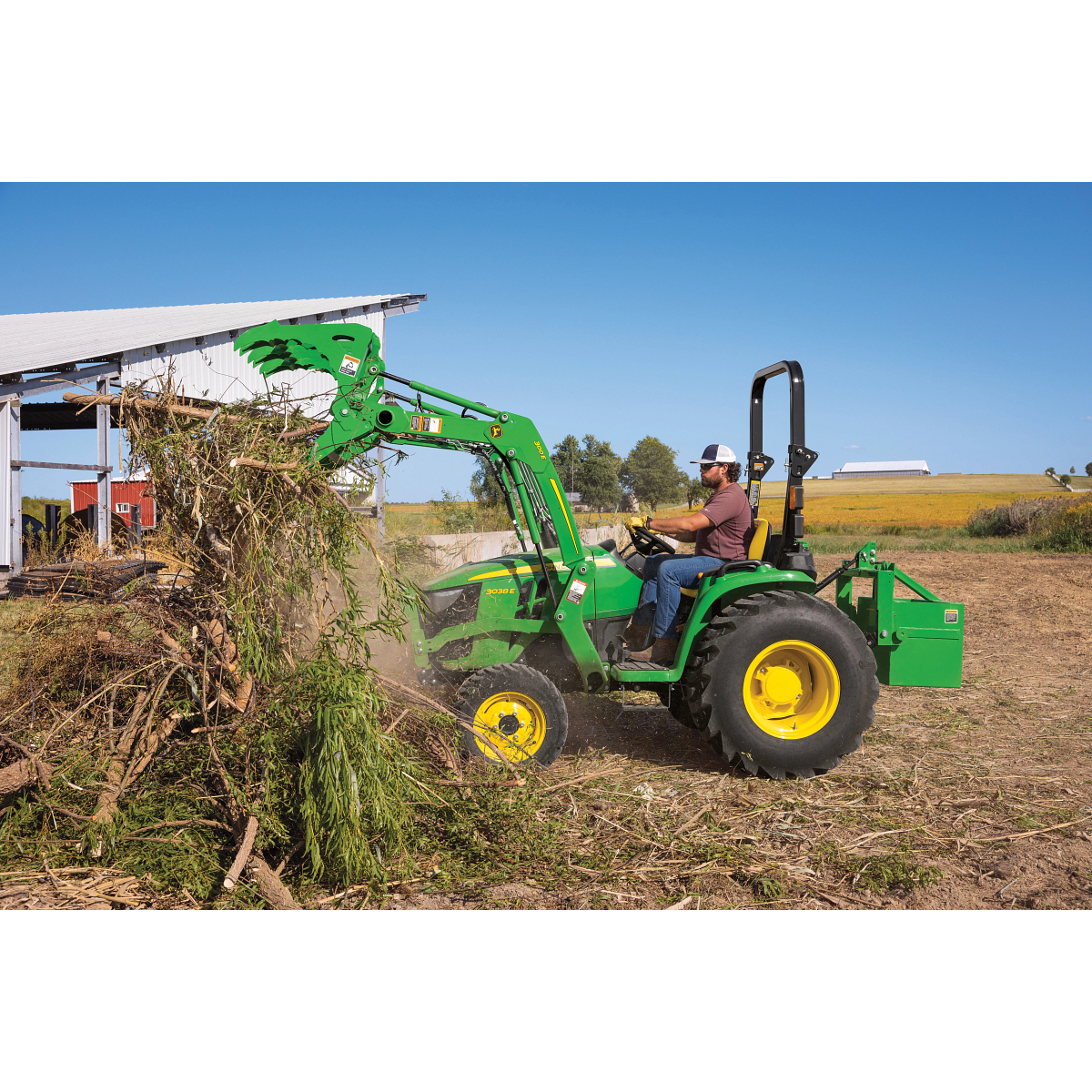Operator in a green tractor with a front loader and grapple lifts a pile of brush near a metal-sided building, with open farmland stretching into the background under a clear blue sky