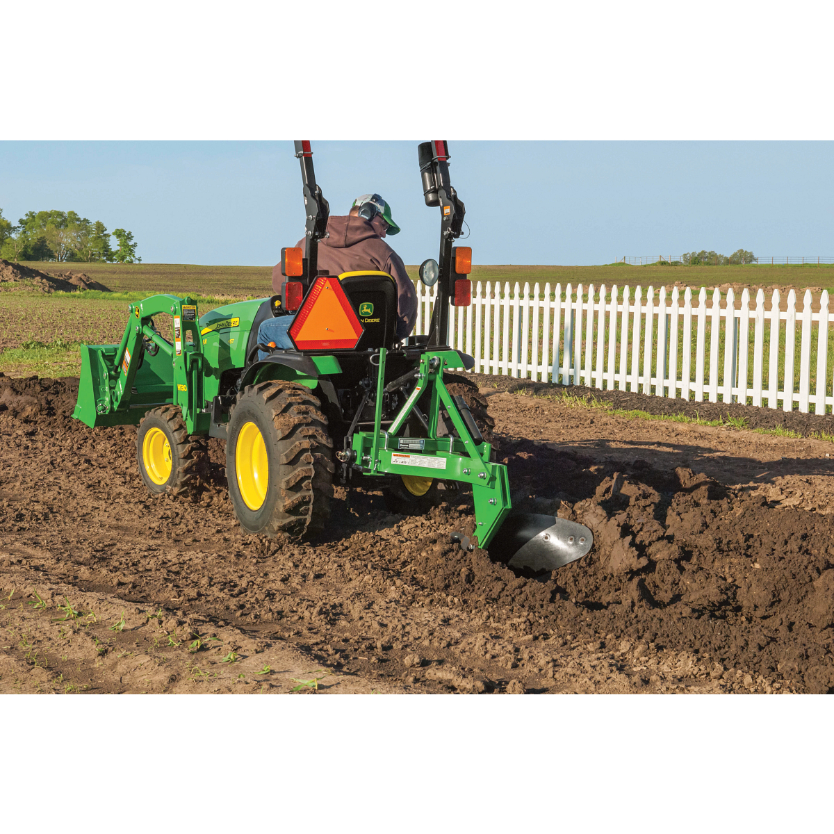 Green tractor with a rear plow attachment cuts a trench through freshly tilled soil near a white picket fence, with a driver seated at the controls and open farmland in the background.