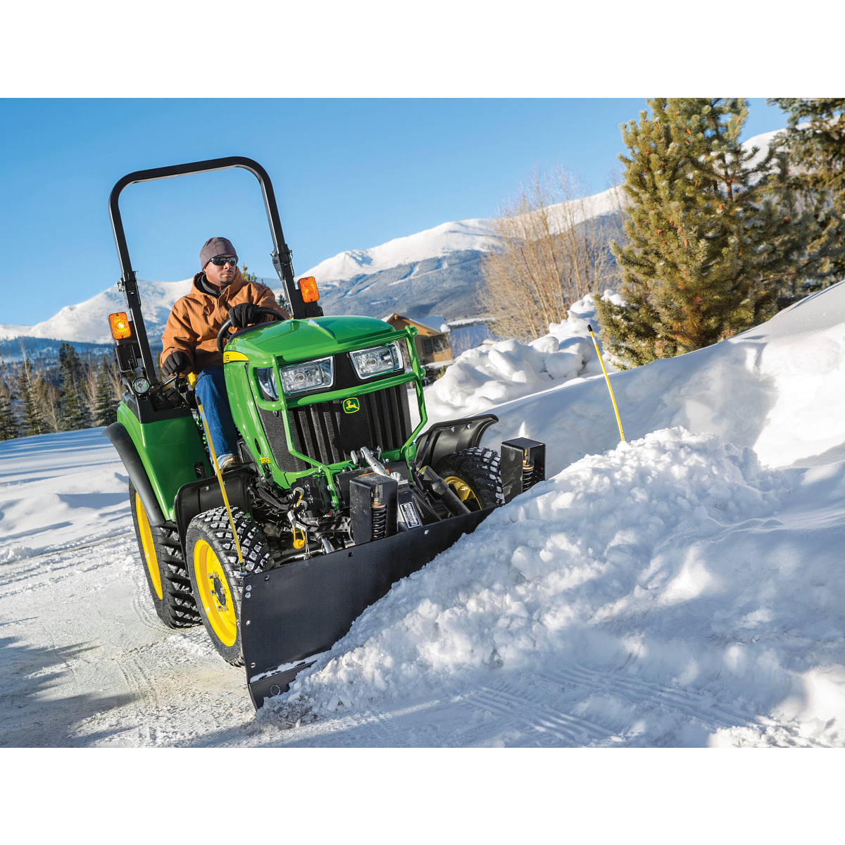 Green tractor with a front-mounted plow clearing snow along a road, pushing a thick ridge of snow to the side with evergreen trees mountains visible in the background under a clear sky