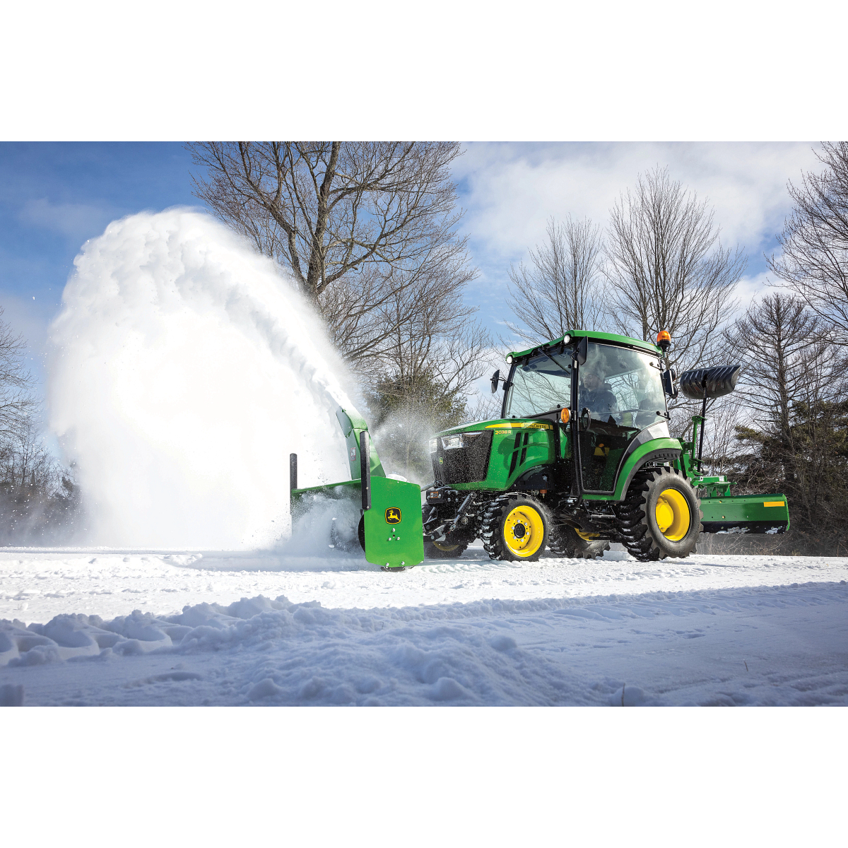 Green tractor equipped with a front-mounted snow blower clearing a snowy path, blasting a high arc of snow to the side next to bare trees and under a cloudy sky