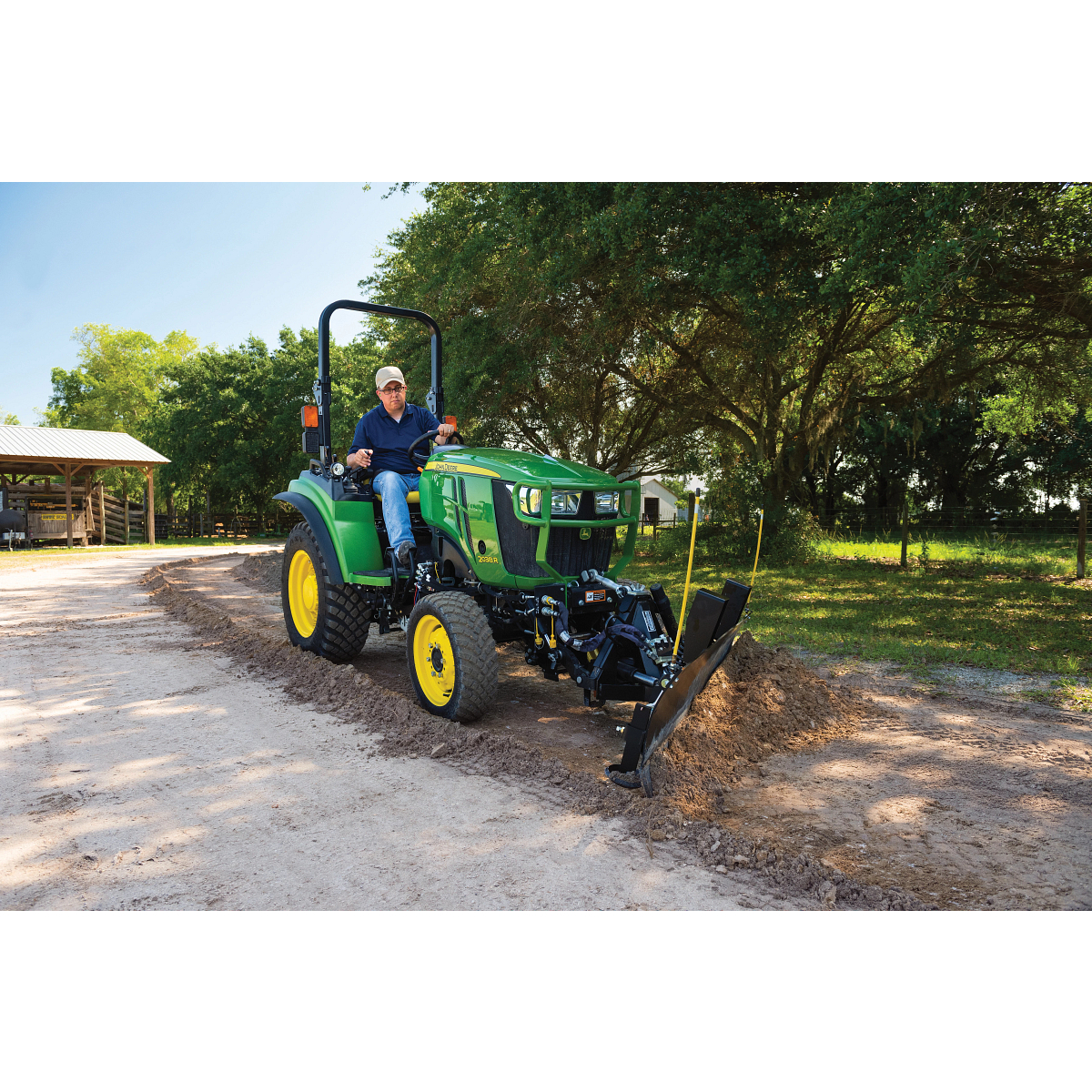 Person operating a compact green tractor with a front attachment leveling a dirt driveway on a rural property with trees, a shed, and dappled sunlight filtering through the foliage