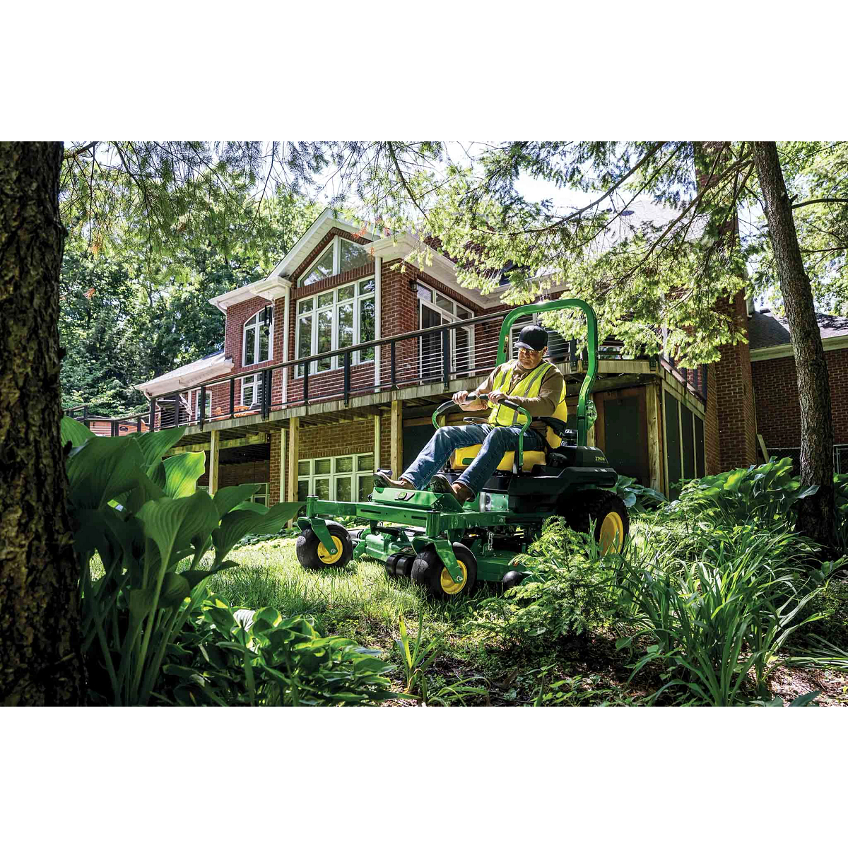 A landscaper mowing grass along the edge of a garden behind a residence with a green zero-turn mower.