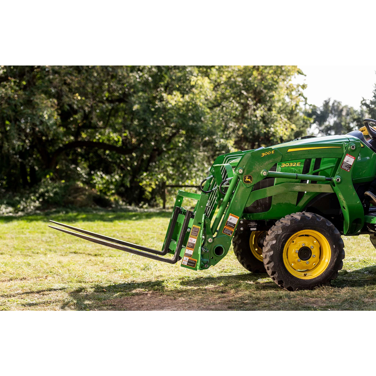 Front left view of the front of a 3032E compact tractor with AP11F pallet fork on a tree-lined lawn