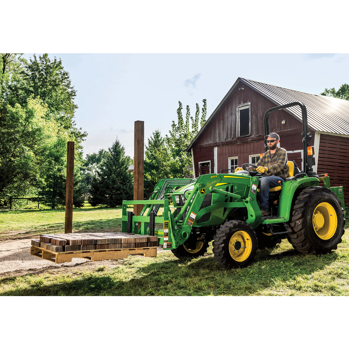 Side left view of a 3032E compact tractor with attached 300E loader and AP11F pallet fork lifting a pallet of brick in front of a barn