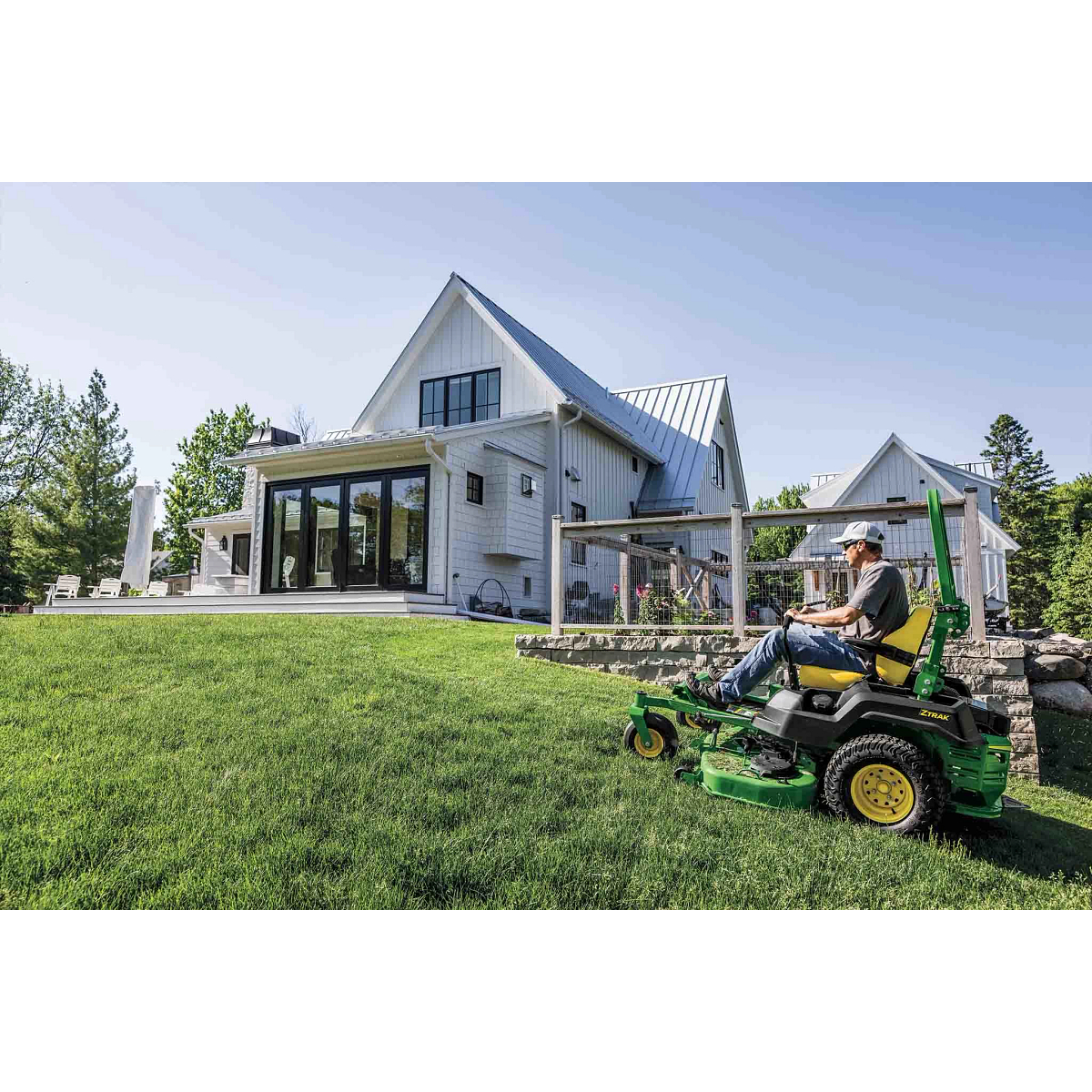 A green zero-turn mower is mowing up a gentle slope near a fenced-off garden.